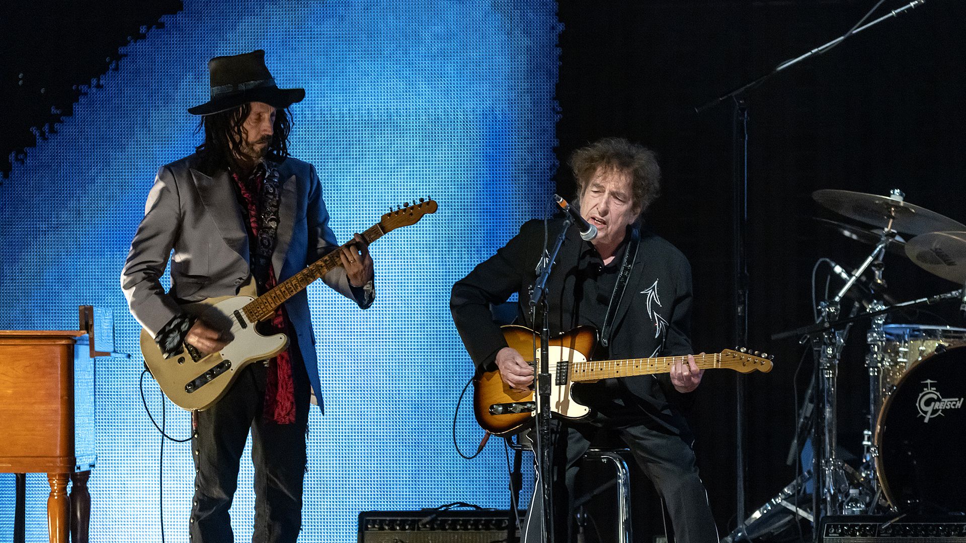 bob dylan on stage with an electric guitar while another man with an instrucment and wearing a hat stands beside him