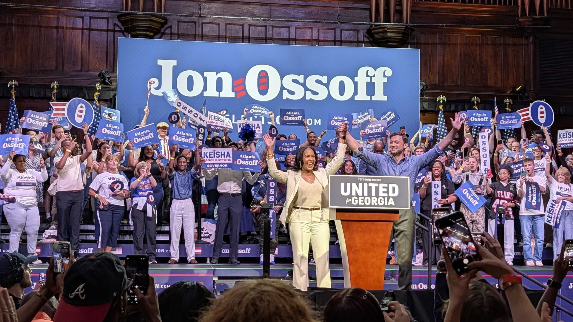 Stage rally with a large blue backdrop reading "Jon Ossoff". Diverse crowd holds blue signs reading "Atlanta for Ossoff"; a woman in a tan suit raises arms  with a man in a blue shirt and green pants at the podium front and center.