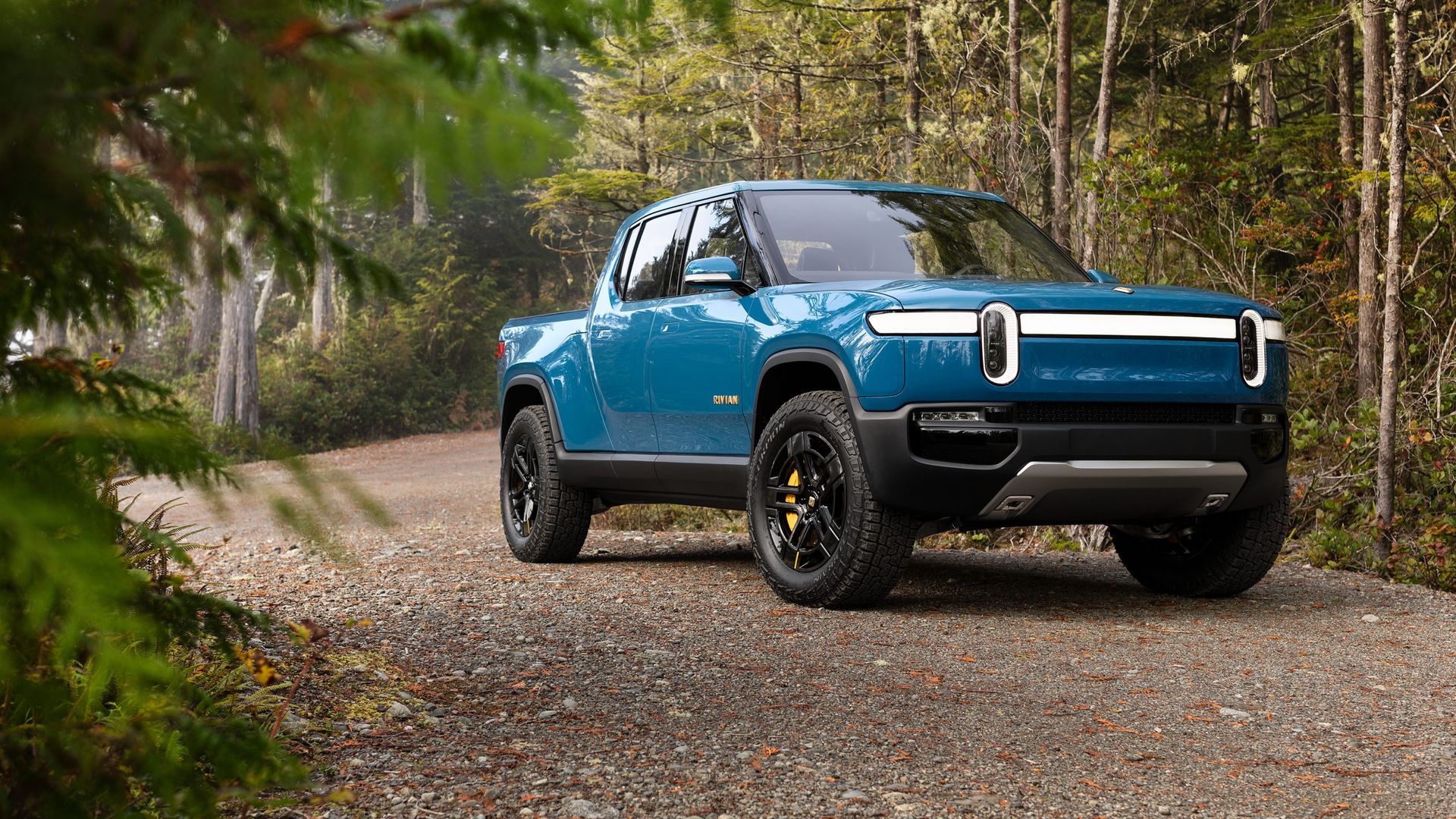Image of a blue Rivian R1T electric truck on a dirt road in the woods. 