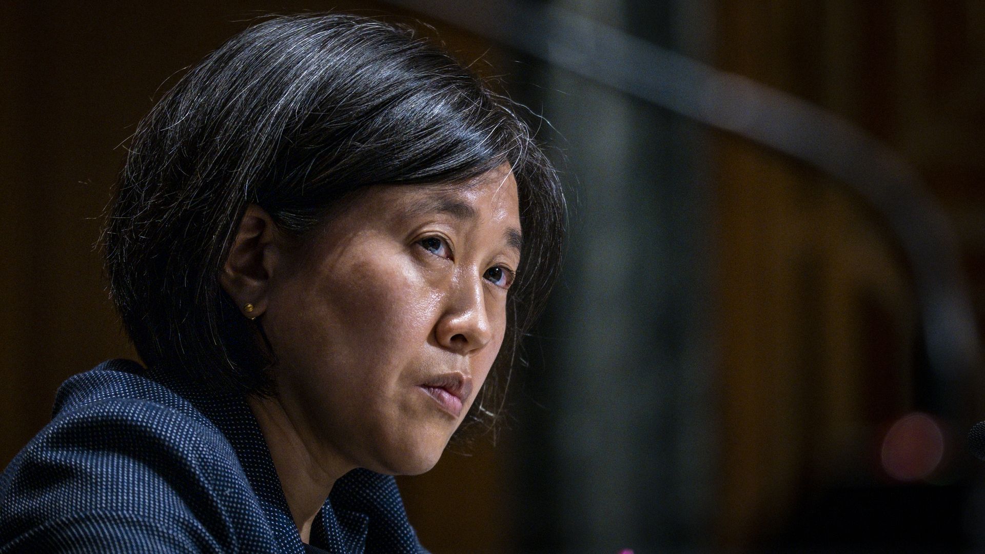 Photo of Katherine Tai in a dark navy blue blazer looking up to her right