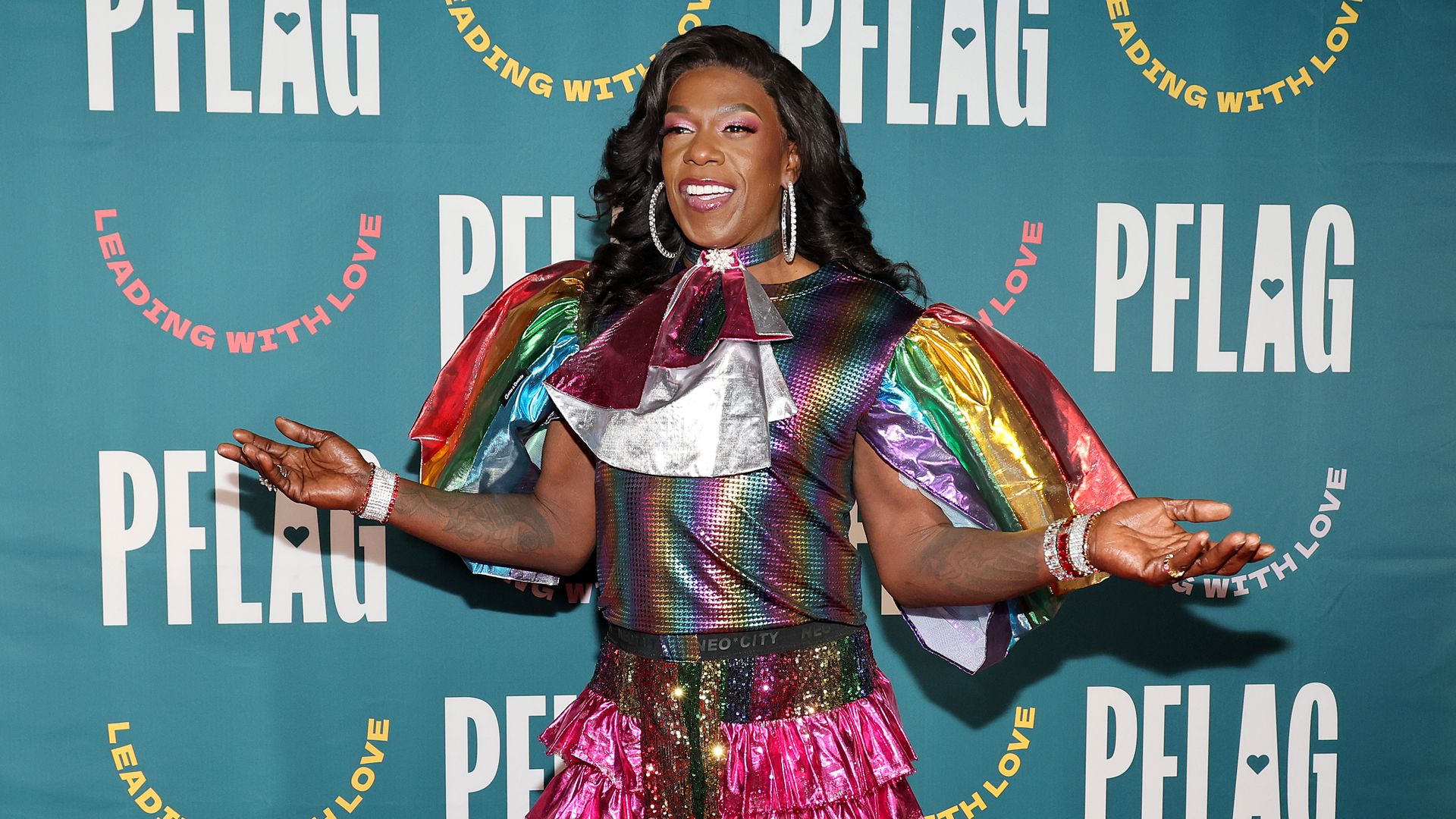 Photo shows Big Freedia smiling with open arms. She has long, dark curly hair and is wearing large diamond hoops and a sparkly rainbow outfit.