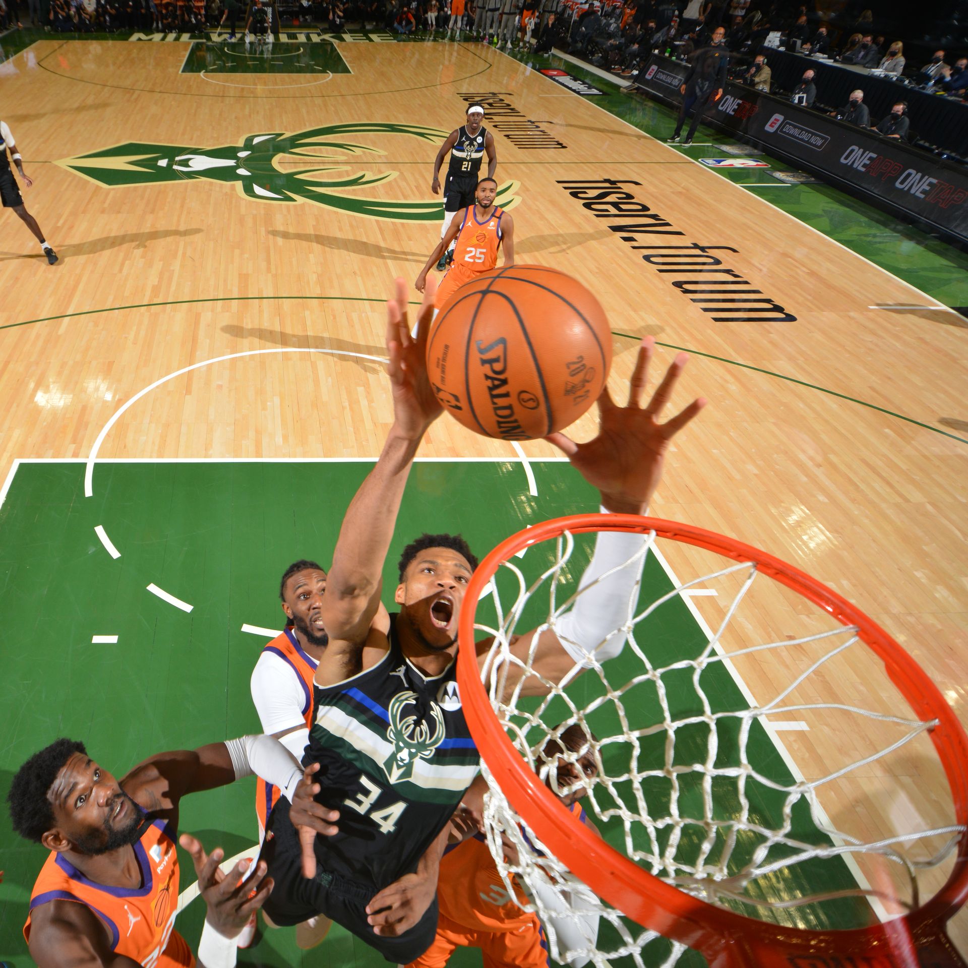 Giannis Antetokounmpo, in a green jersey, #34 of the Milwaukee Bucks, jumps to dunk a basketball into a hoop above Phoenix Suns players in orange uniforms.