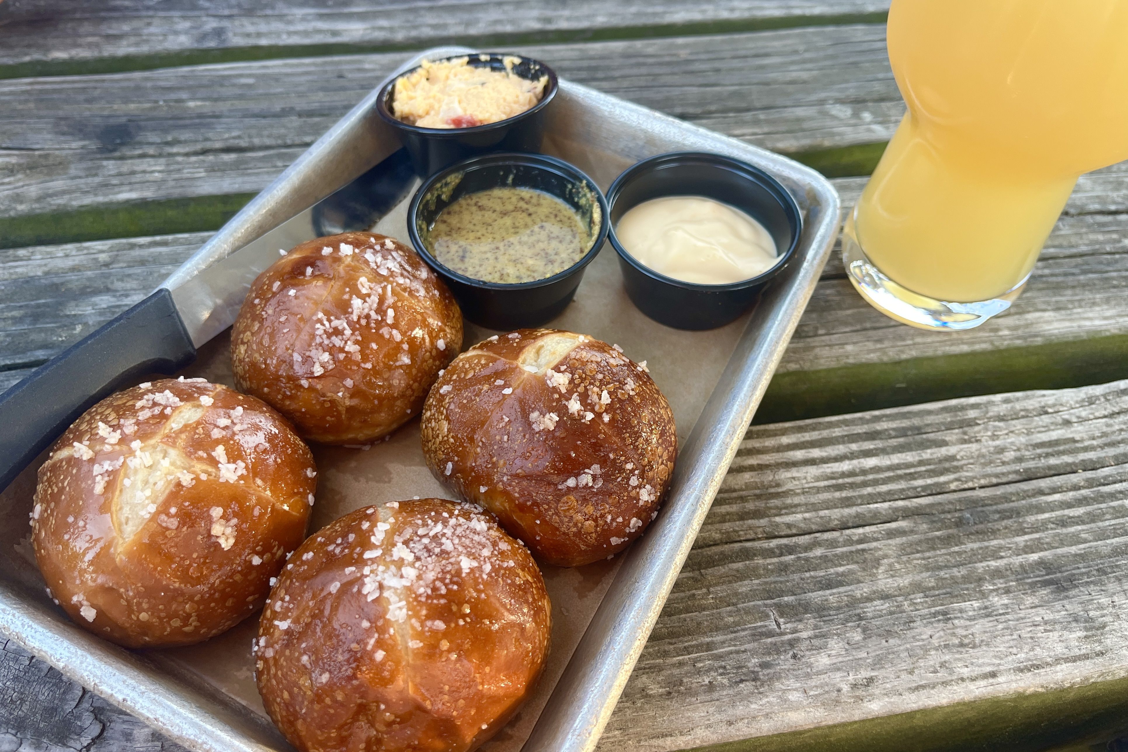 Tray of glossy pretzel buns with coarse salt on a metal tray, three black dipping cups (mustard, creamy sauce, orange-tinted dip) and a glass of orange juice on a weathered wooden table.