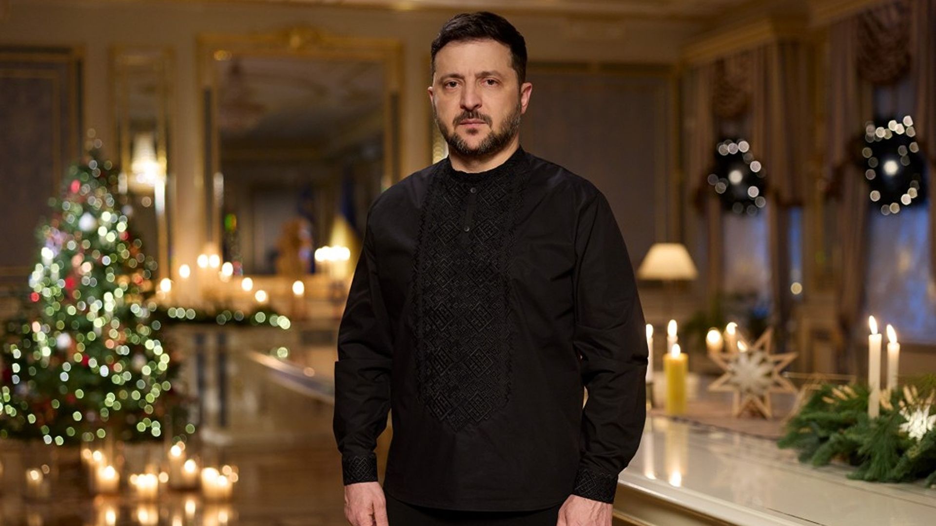 A fair-skinned Ukrainian President Volodymyr Zelensky, who has short, dark hair and is wearing a black fleece top and black pants, looks intently with his dark brown eyes into the camera in front of lit white candles (R) and a Christmas tree that's decorated with yellow lights and a few red baubles.