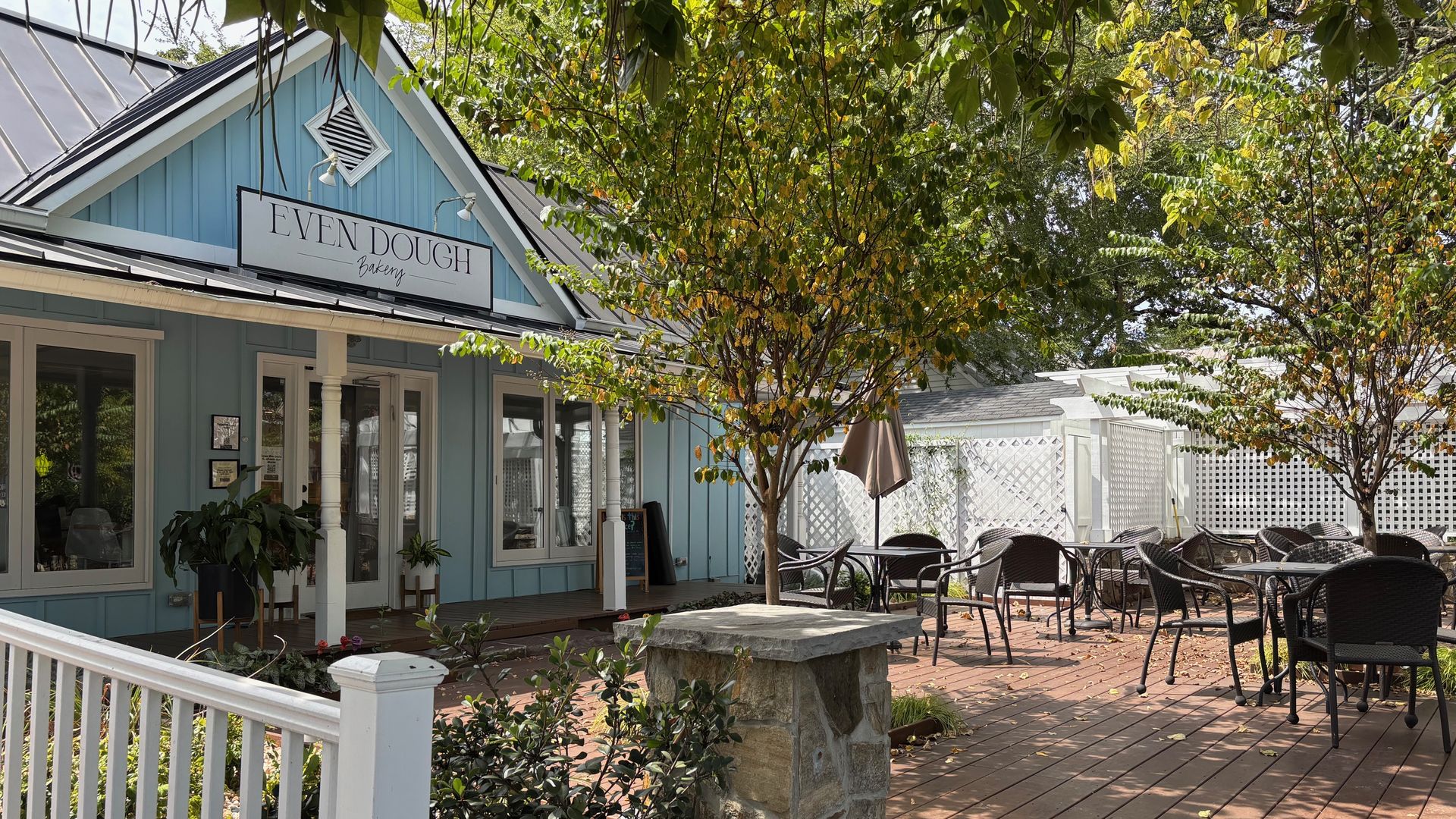 Outdoor patio seating with black chairs and tables under trees with green leaves beside a light blue building with a sign reading "EVEN DOUGH Bakery" and white railing in front.
