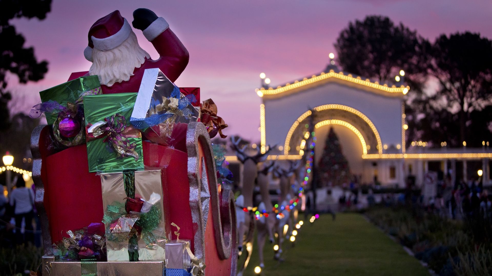 Santa in his sleigh full of presents facing Spreckels Organ Pavilion lit up with Christmas lights in Balboa Park.