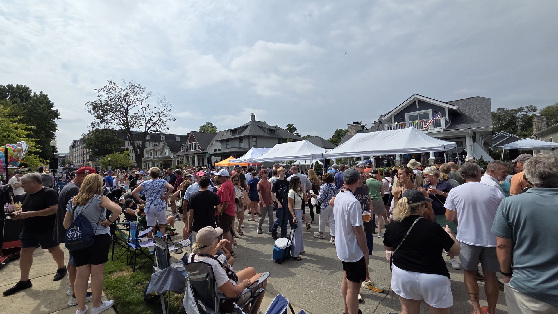 Crowd at a street festival in a small town. White vendor tents line the street, colorful balloons on the left, houses with peaked roofs in the background, people chatting, strolling, and drinking.
