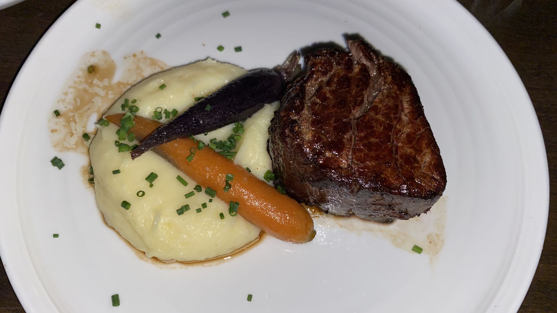 A piece of steak on a round white plate next to two carrots and a pile of mashed potatoes. 