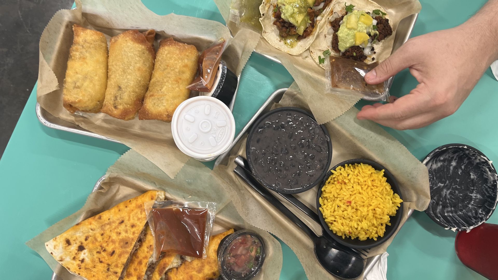 Three golden-brown fried egg rolls on parchment, two tacos with green and yellow toppings with a hand holding a sauce packet, yellow rice and black beans in black bowls, and quesadilla slices with salsa.