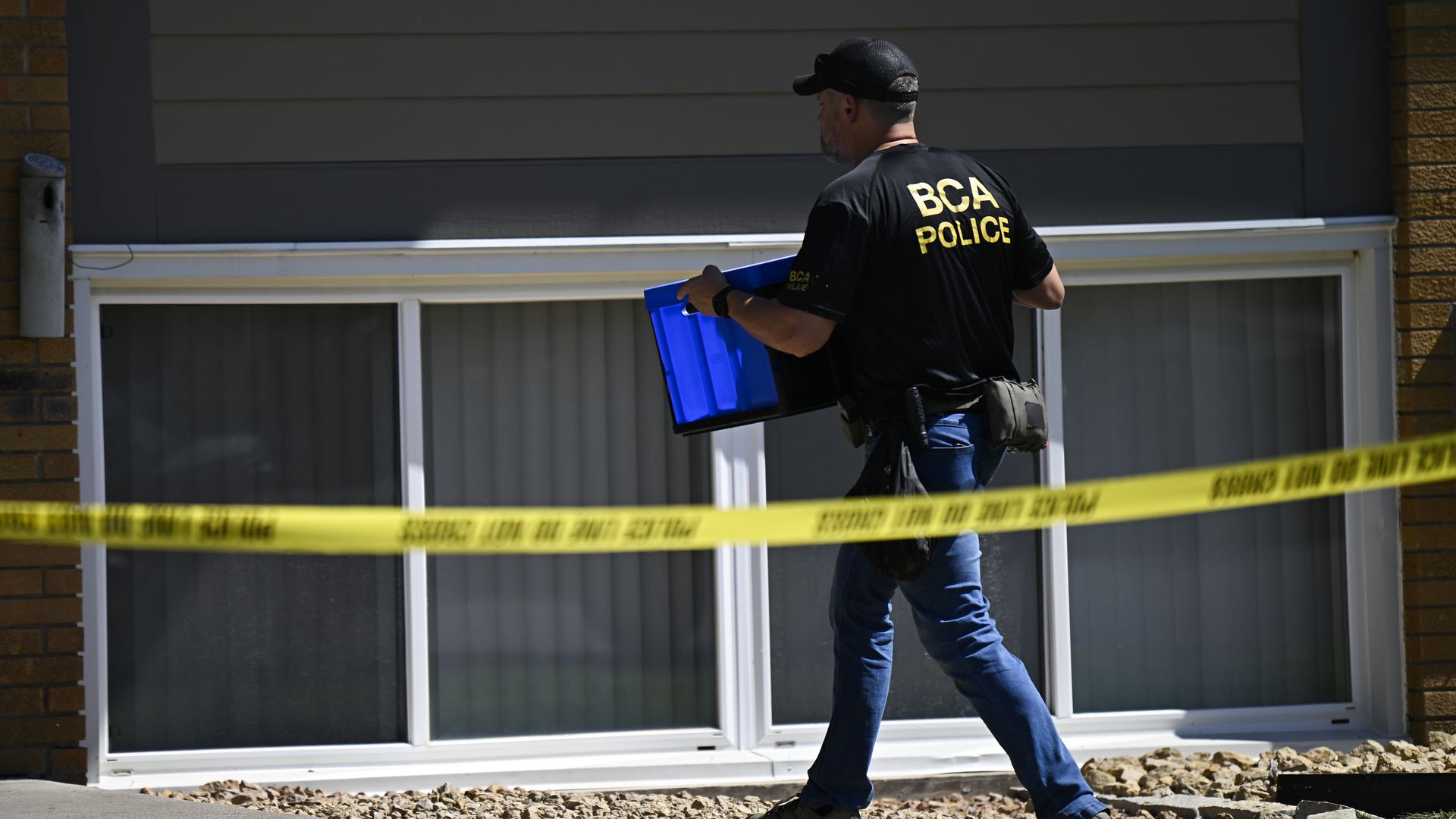 A BCA police officer in black shirt and blue jeans carries a blue bin past yellow police tape near a building with large windows and brown brick walls on a sunny day.
