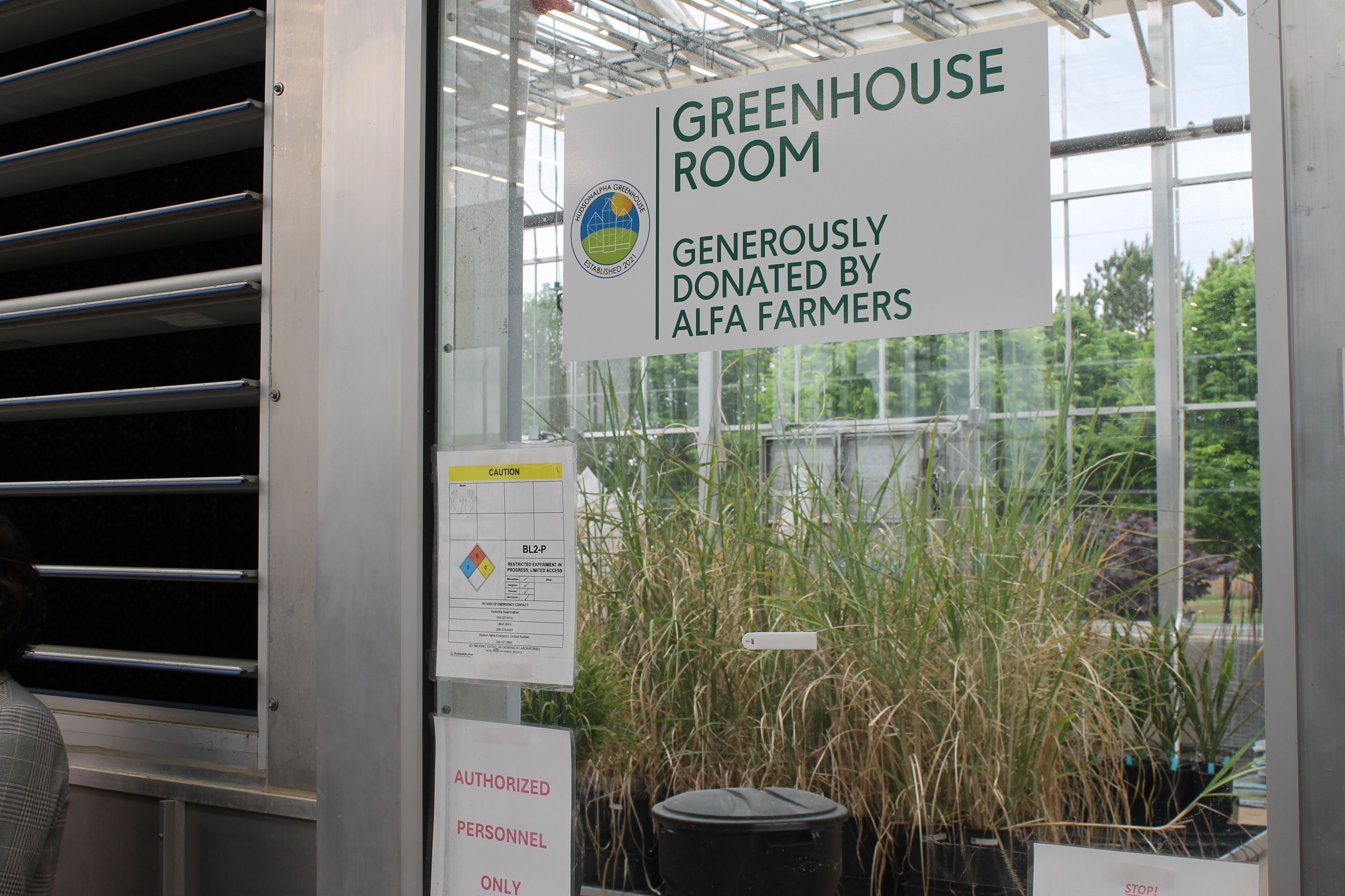 Sign on glass wall reads "Greenhouse Room" in green letters; a circular logo and the text "Generously donated by Alfa Farmers." Metal frame, glass, and tall plants outside.