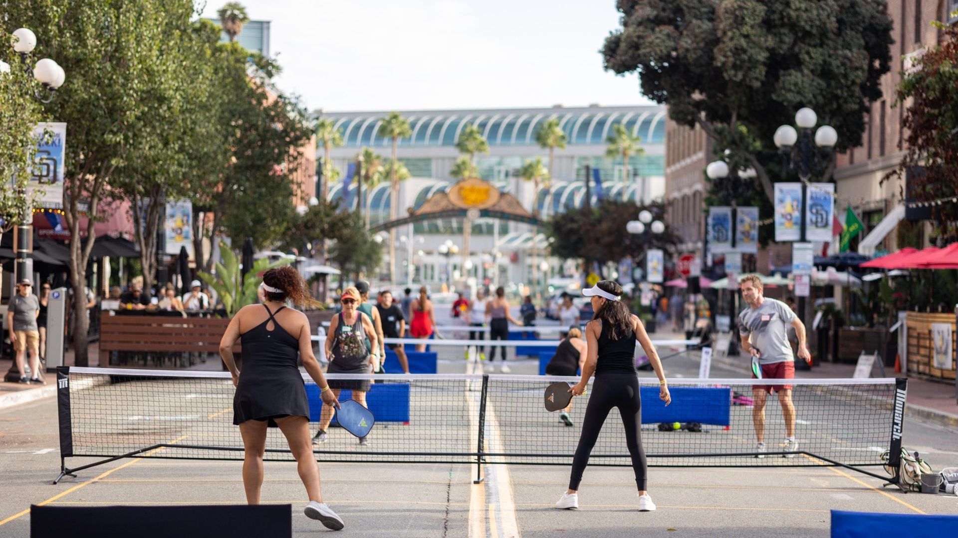 Street converted to a pickleball court with nets; players in athletic wear hold paddles as spectators line the urban plaza, palm trees, banners, and a glass-curved building in the background.