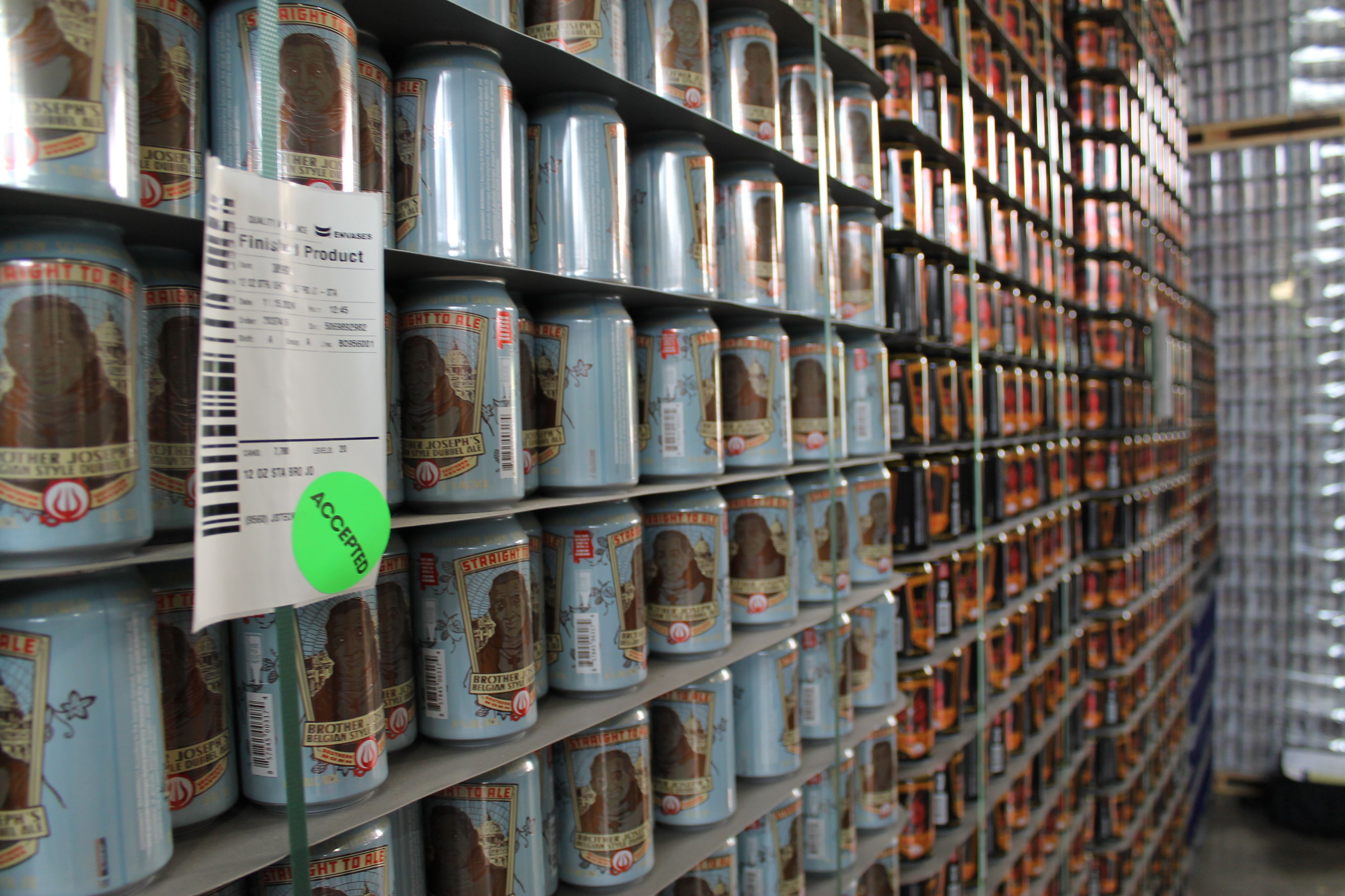 Rows of cans labeled "Brother Joseph's Belgian Style Dubbel Ale" and other beer cans stacked on shelves in a warehouse, with a green "ACCEPTED" sticker on a label attached to one shelf.