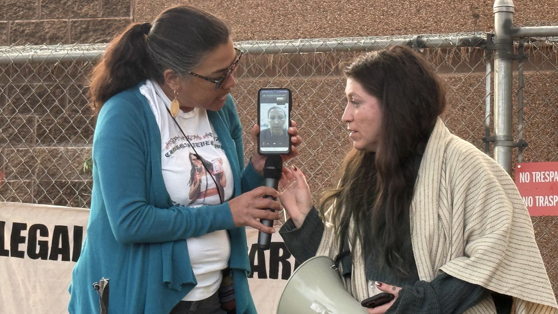 Two women stand while holding a smartphone showing a person on video call.