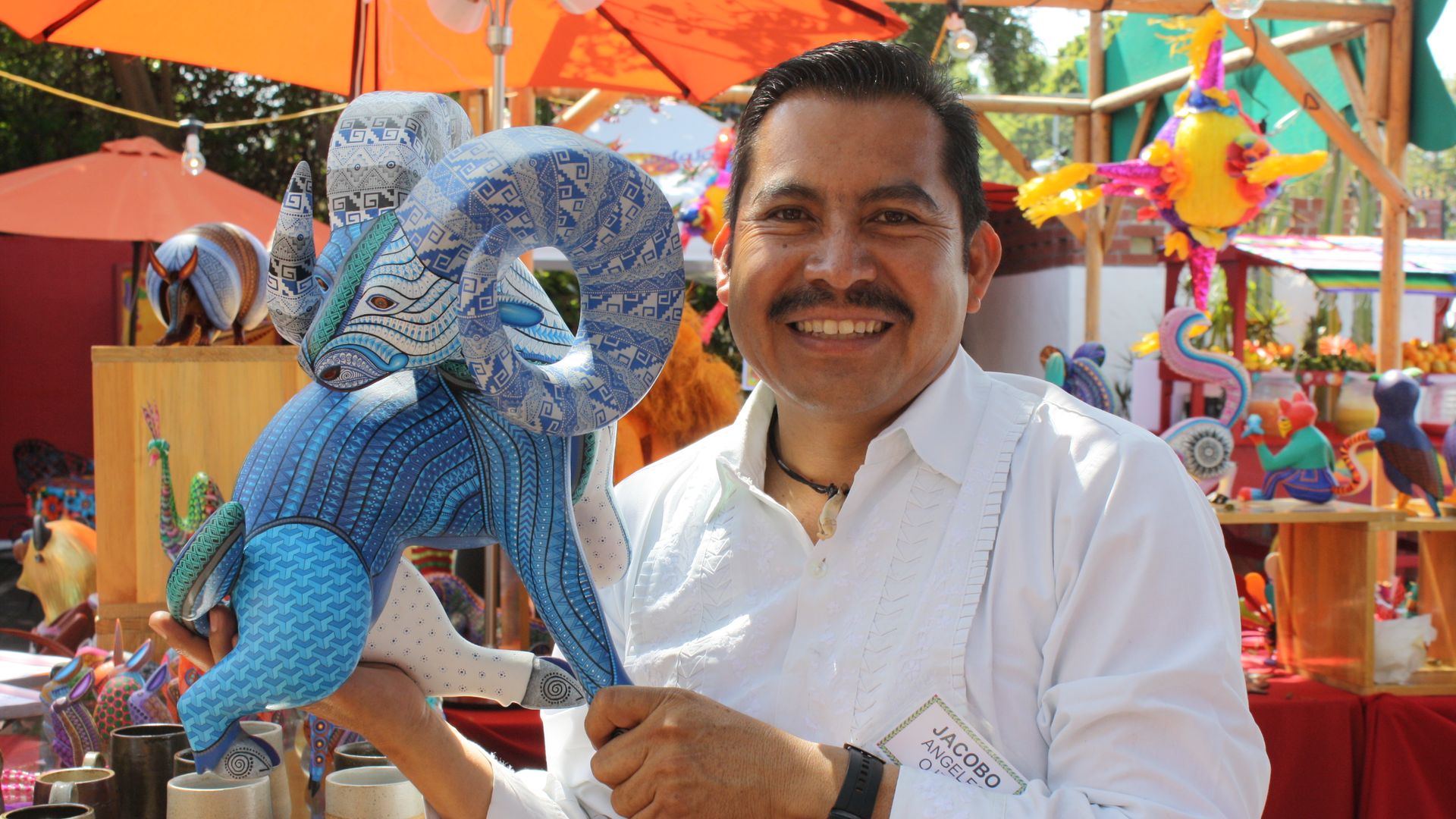 Man in white shirt smiles holding a detailed blue ram sculpture at an outdoor artisan market with colorful crafts and orange umbrellas in the background.