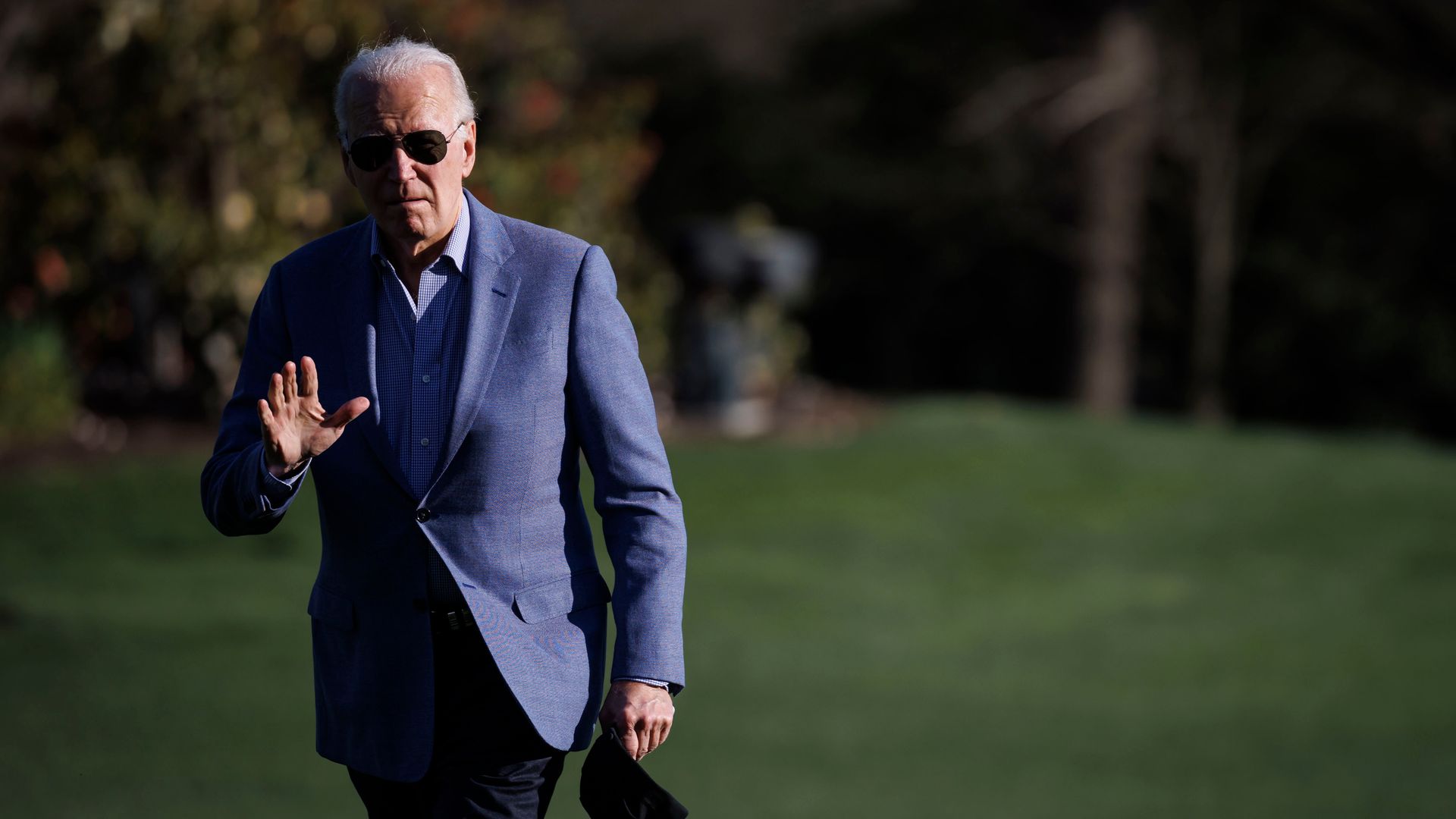 President Biden, dressed in slacks and a sports coat, waves at a camera while walking across a lawn.