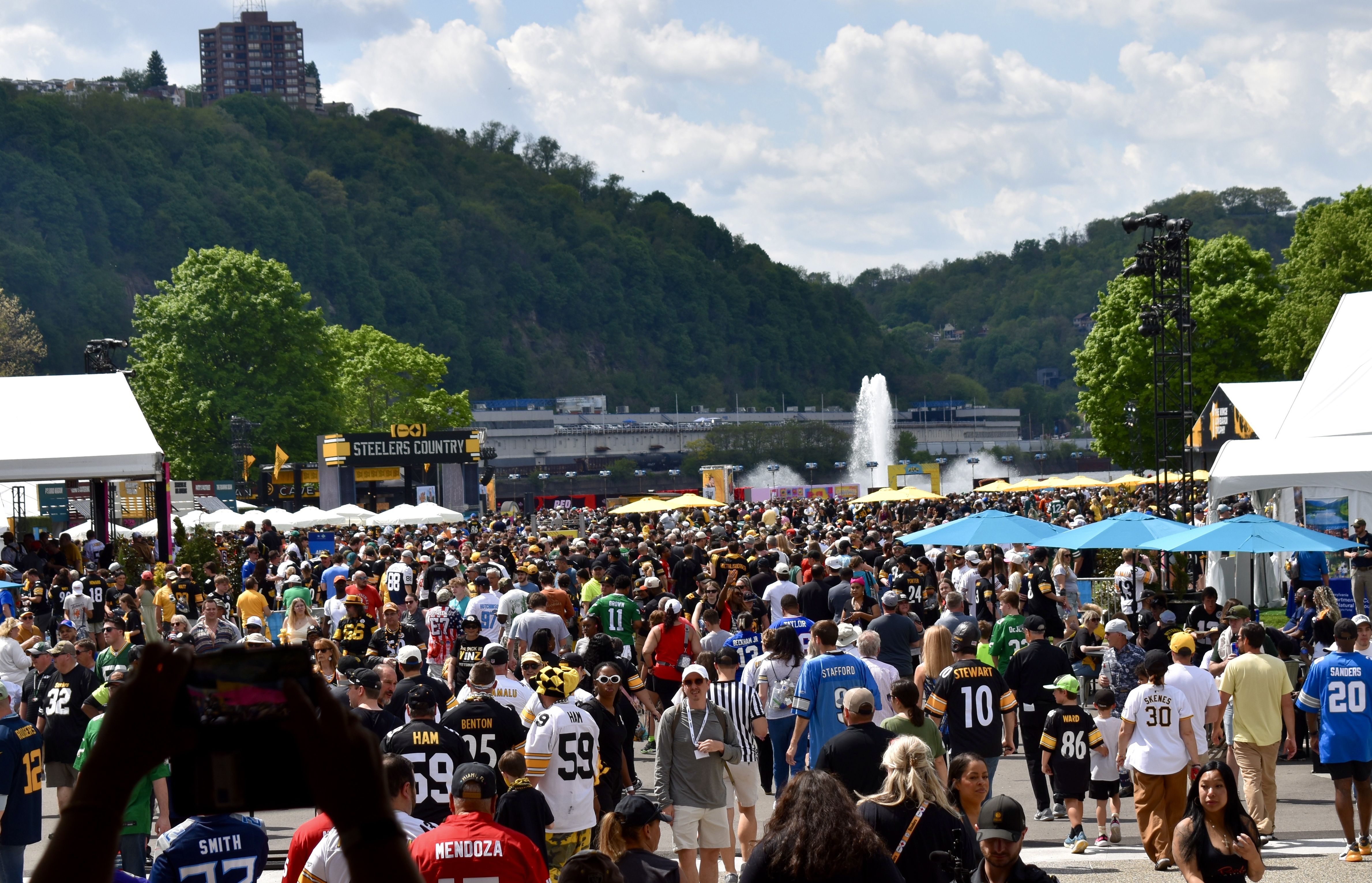 Massive outdoor festival with hundreds of fans in Steelers jerseys beneath white tents and yellow umbrellas; a "STEELERS COUNTRY" sign, distant hills, a fountain, and tall stage lights on the right.
