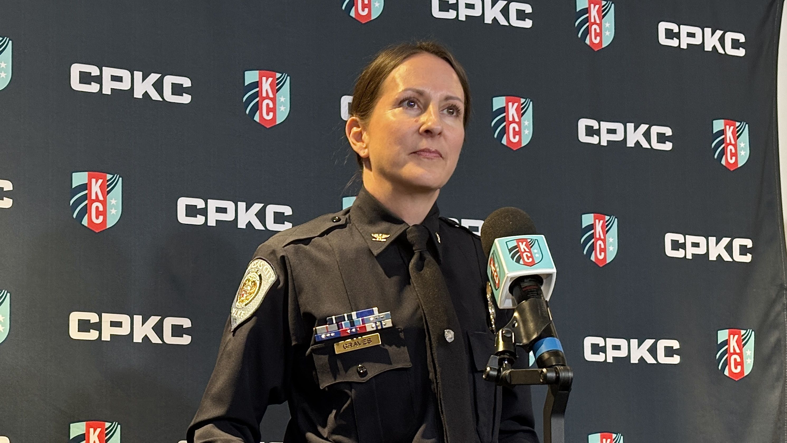 Woman police officer in black uniform with KC police badge at podium speaking in front of a backdrop featuring CPKC logos and KC shield emblem in red, teal, white.