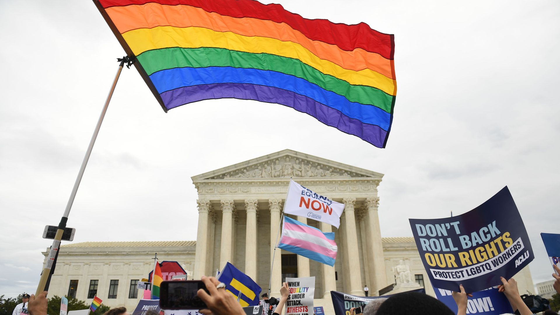 In this image, an LGBTQ rainbow flag flies above the Supreme Court while protestors hold signs that read: "Don't roll back our rights, protect LGBTQ workers"