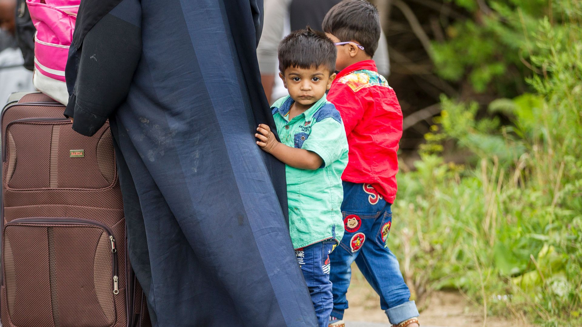 A little boy stands holding onto an adult's clothing.