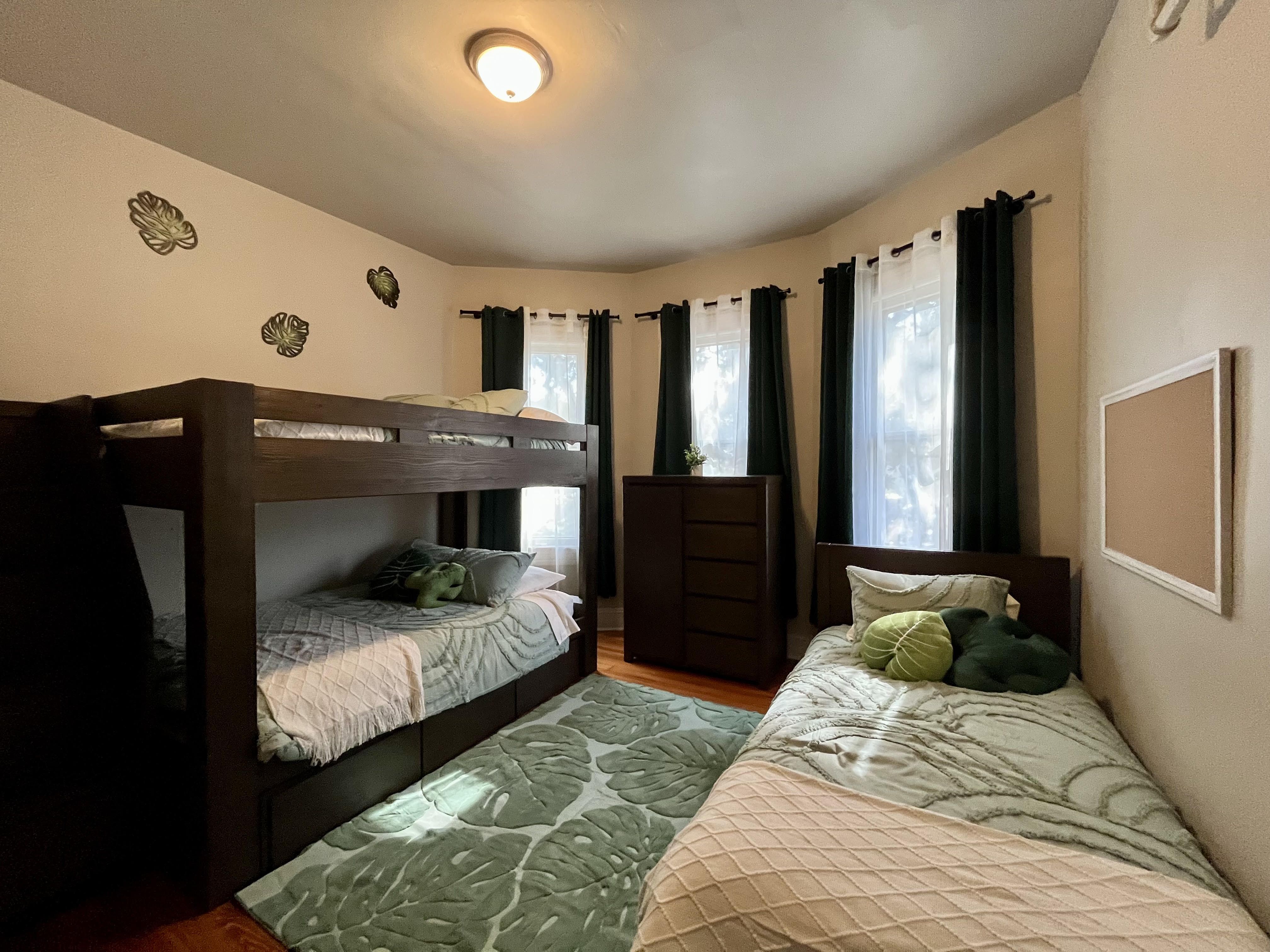 A bedroom inside the new 12-bed crisis nursery house in San Antonio has a dark wood bunk bed and a twin bed, with green leaf-patterned quilts, beige walls with leaf decals, and large windows.