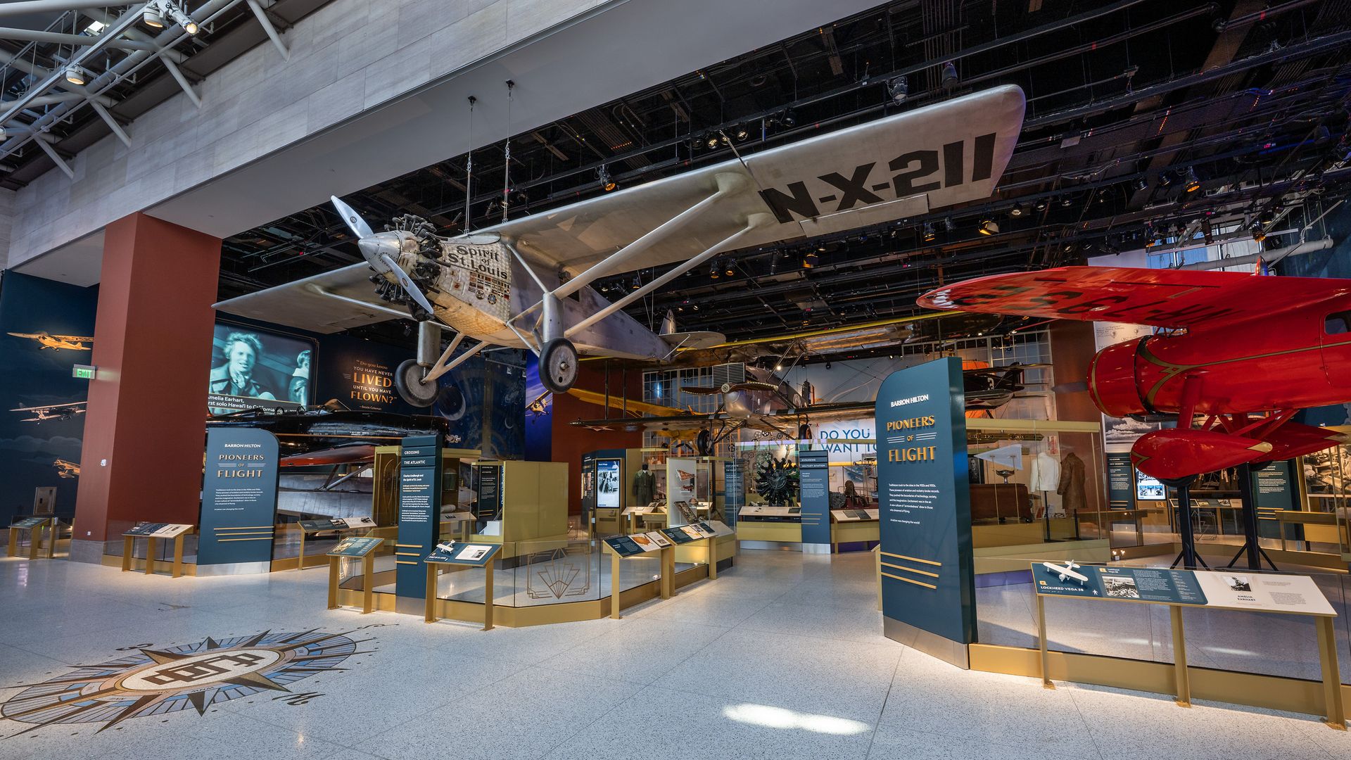 Museum exhibit titled "Pioneers of Flight" with vintage planes including the Spirit of St. Louis, surrounded by informational displays and a tiled compass on the floor.