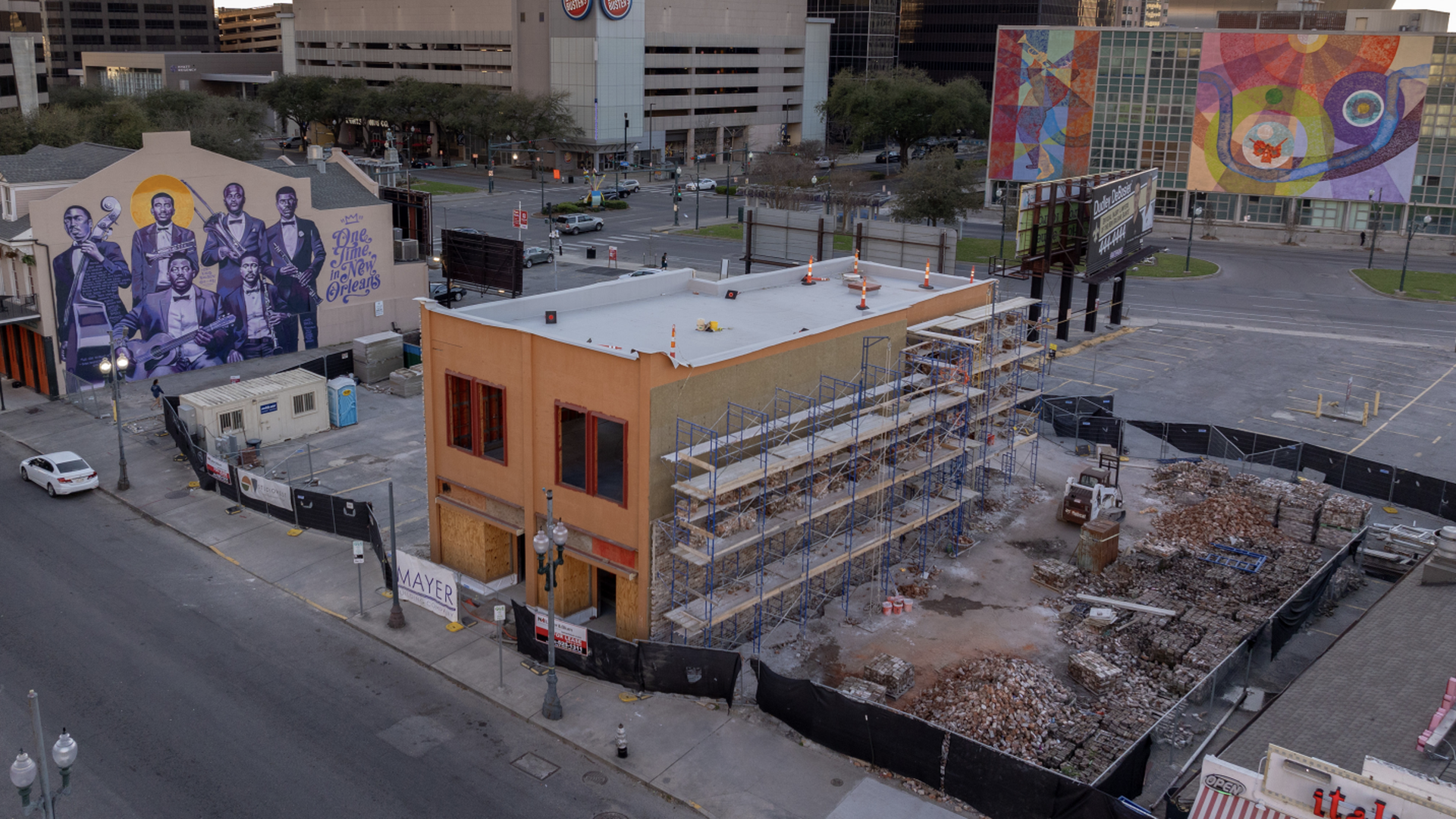Construction site with a building under renovation surrounded by scaffolding and debris, mural of jazz musicians on one side, colorful abstract murals on a distant building, urban street setting.
