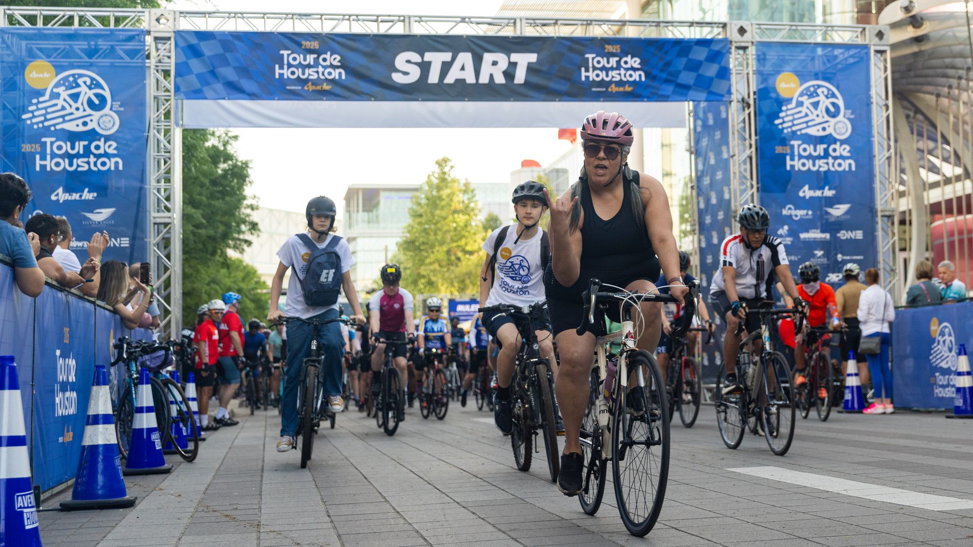 Cyclists of various ages and attire start the 2025 Tour de Houston bike race under a blue banner marked START, with spectators and trees lining the paved urban route.