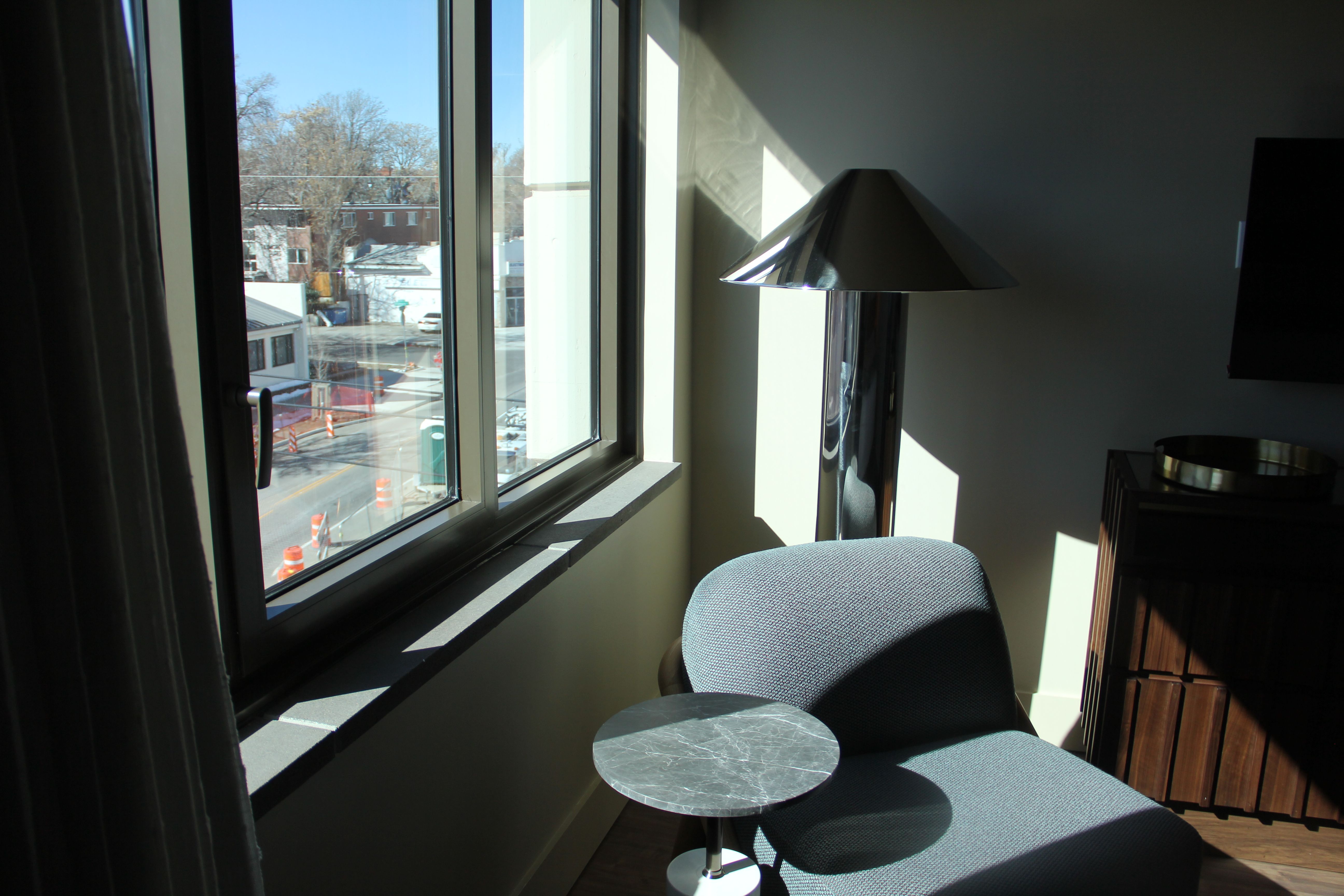 Sunlit hotel room corner with a gray-blue upholstered chaise, a small round marble-topped table, and a tall chrome lamp by a large window; outside, a street with orange traffic cones and bare trees.