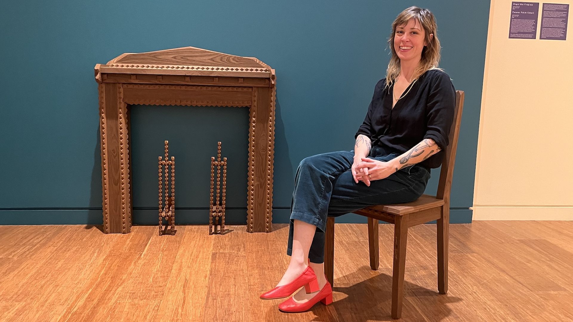 Andi Crist with tattoos in black top, dark pants, and red shoes sits on a wooden chair smiling in front of a carved wooden fireplace frame against a teal wall in a gallery.