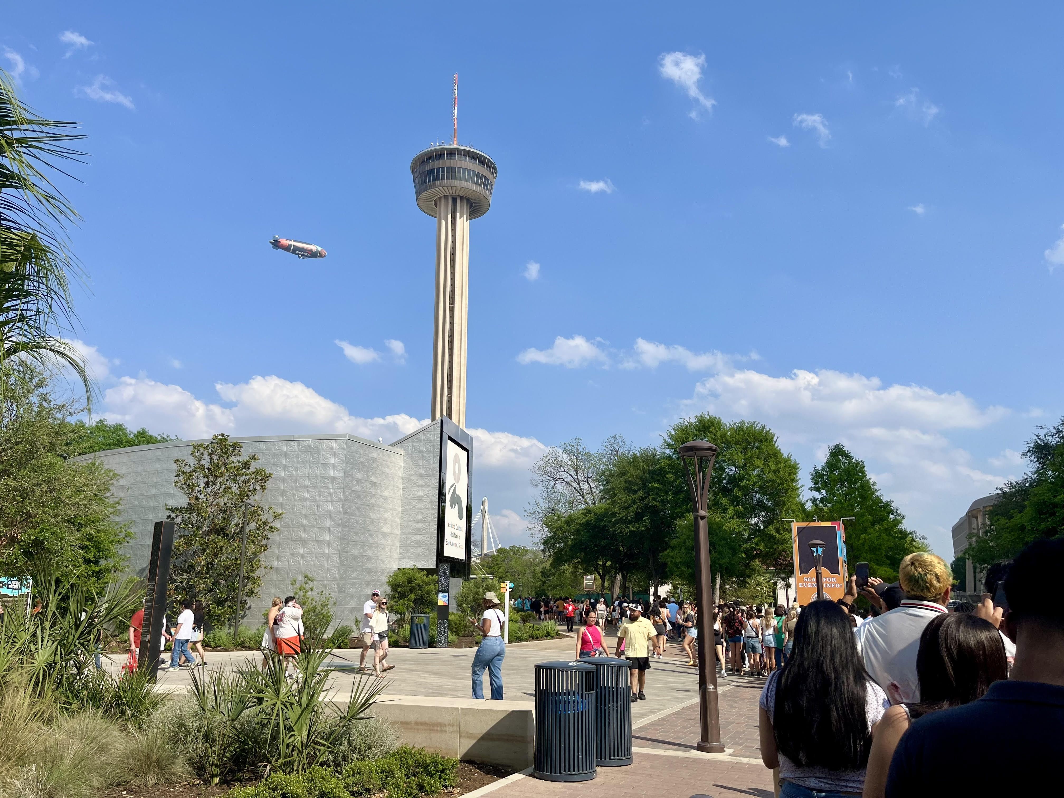 A line wraps around to get into the music festival, with the Tower of the Americas and a blimp seen in the background.