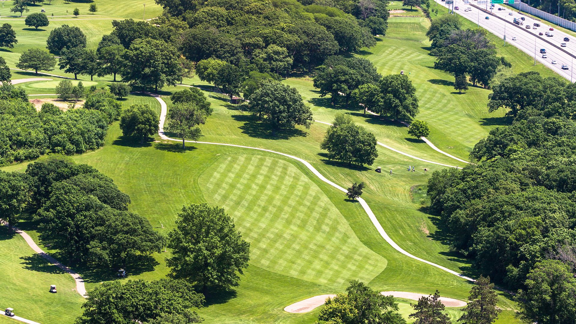 Aerial view of a green golf course with striped fairways, surrounded by dense trees, with a busy highway and city skyline under a clear blue sky in the background.