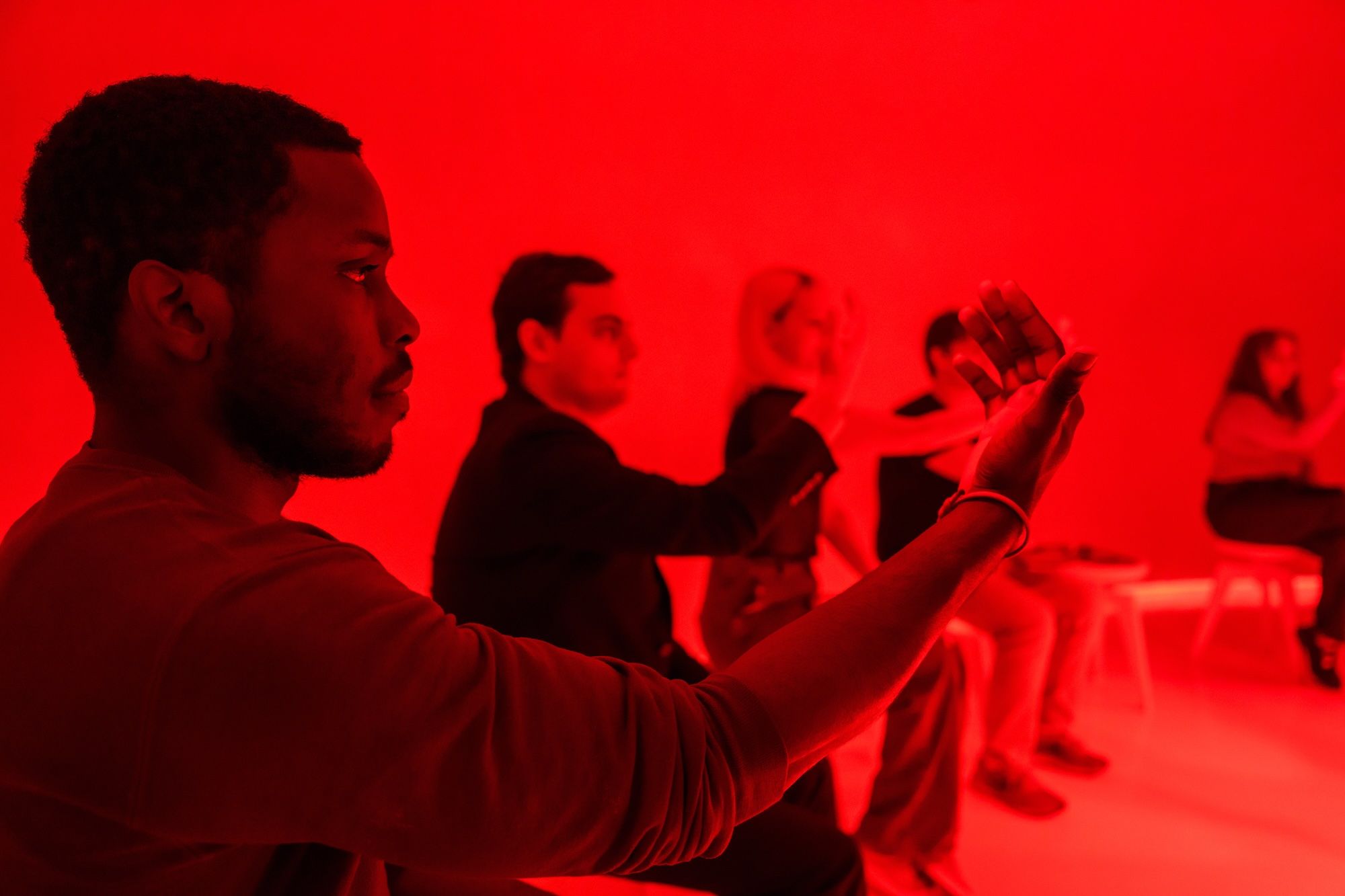 A man in the foreground in profile, holding up a smartphone to take a photo, with several people standing in a red-lit room in the background.