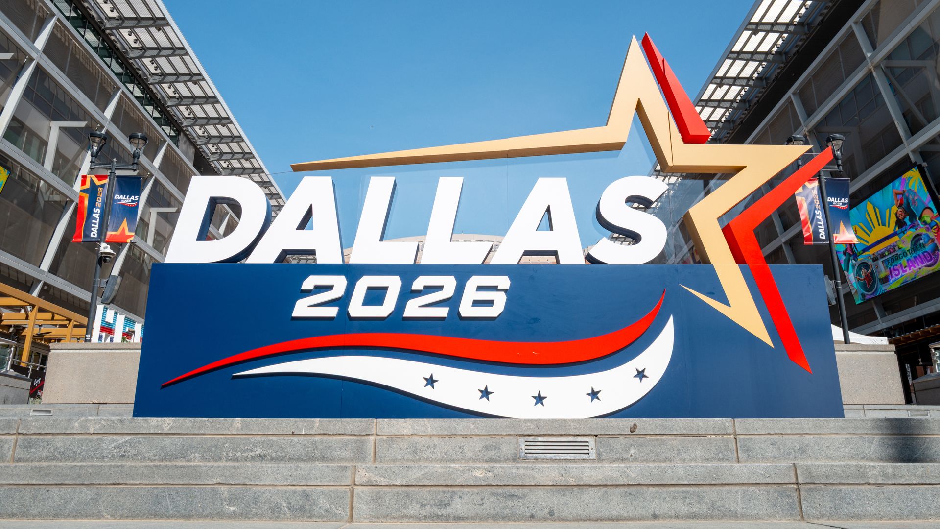Bright blue sky over a large display reading "DALLAS 2026" with a gold and red star graphic; modern glass buildings flank the entrance, colorful banners, and concrete steps.