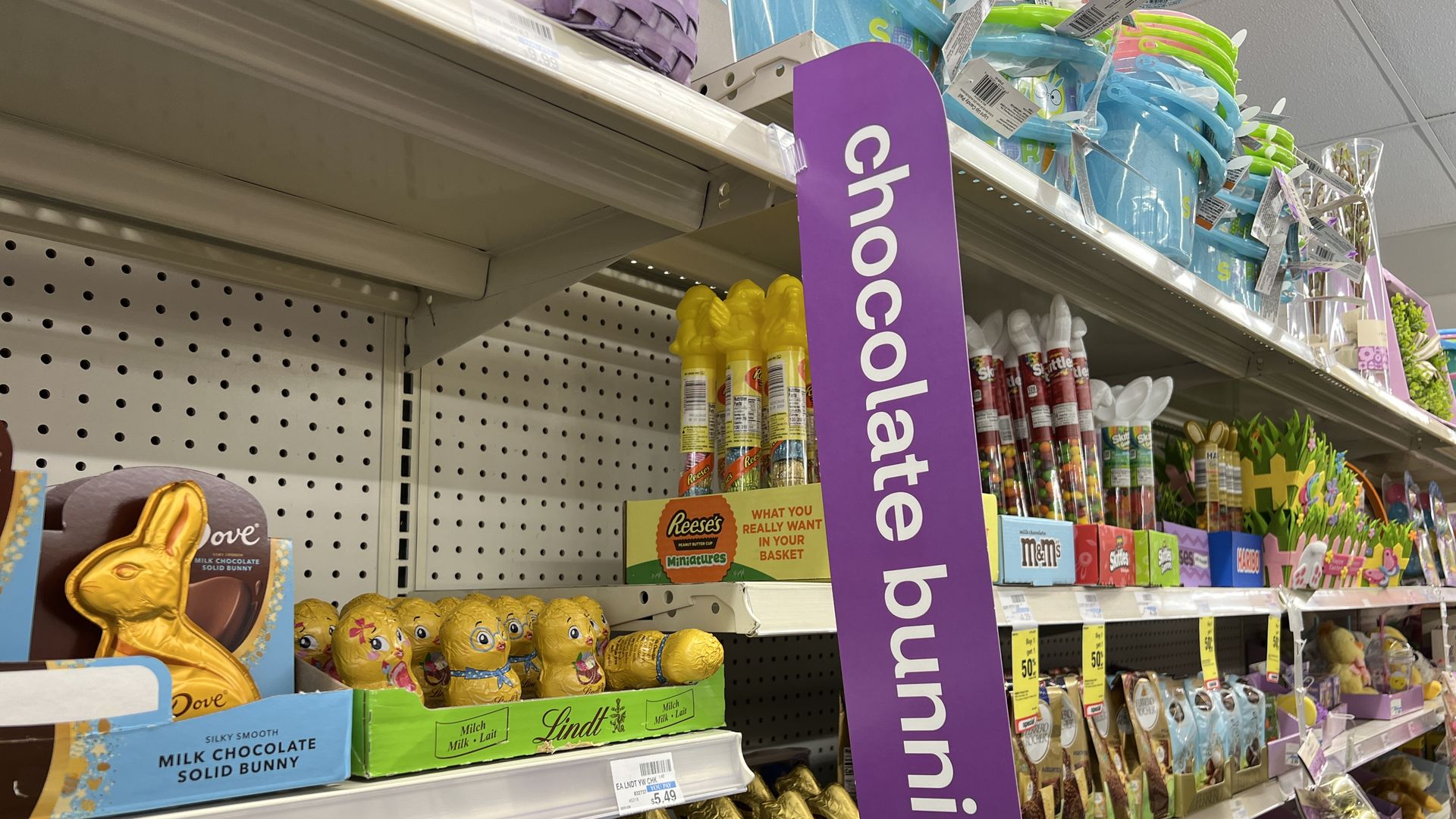 Store shelf of Easter candy featuring a tall purple chocolate bunnies sign; shelves hold bunnies, Lindt eggs, Reese's minis, and other treats in bright packaging.
