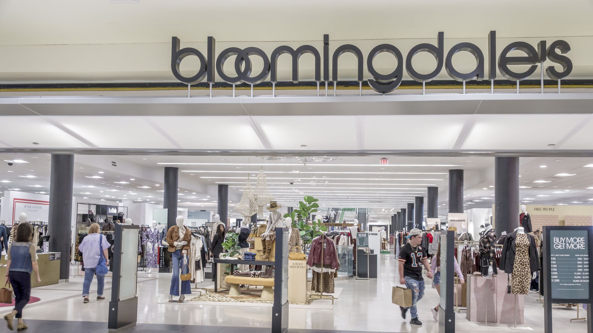 Inside a bright mall, a large Bloomingdale's sign in black letters hangs above a bustling clothing store with mannequins, racks of fashion, and shoppers walking amid displays.