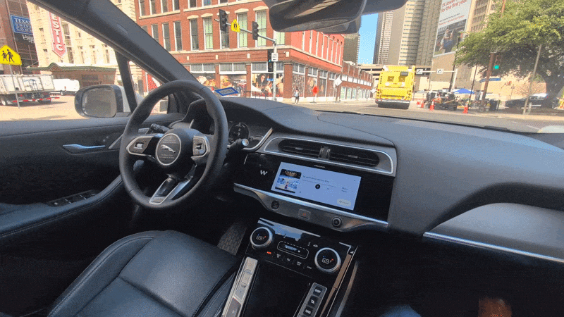 Inside a modern car, front seats visible, steering wheel with Jaguar badge, large central touchscreen, climate controls below; city street with brick buildings and a yellow ambulance outside.