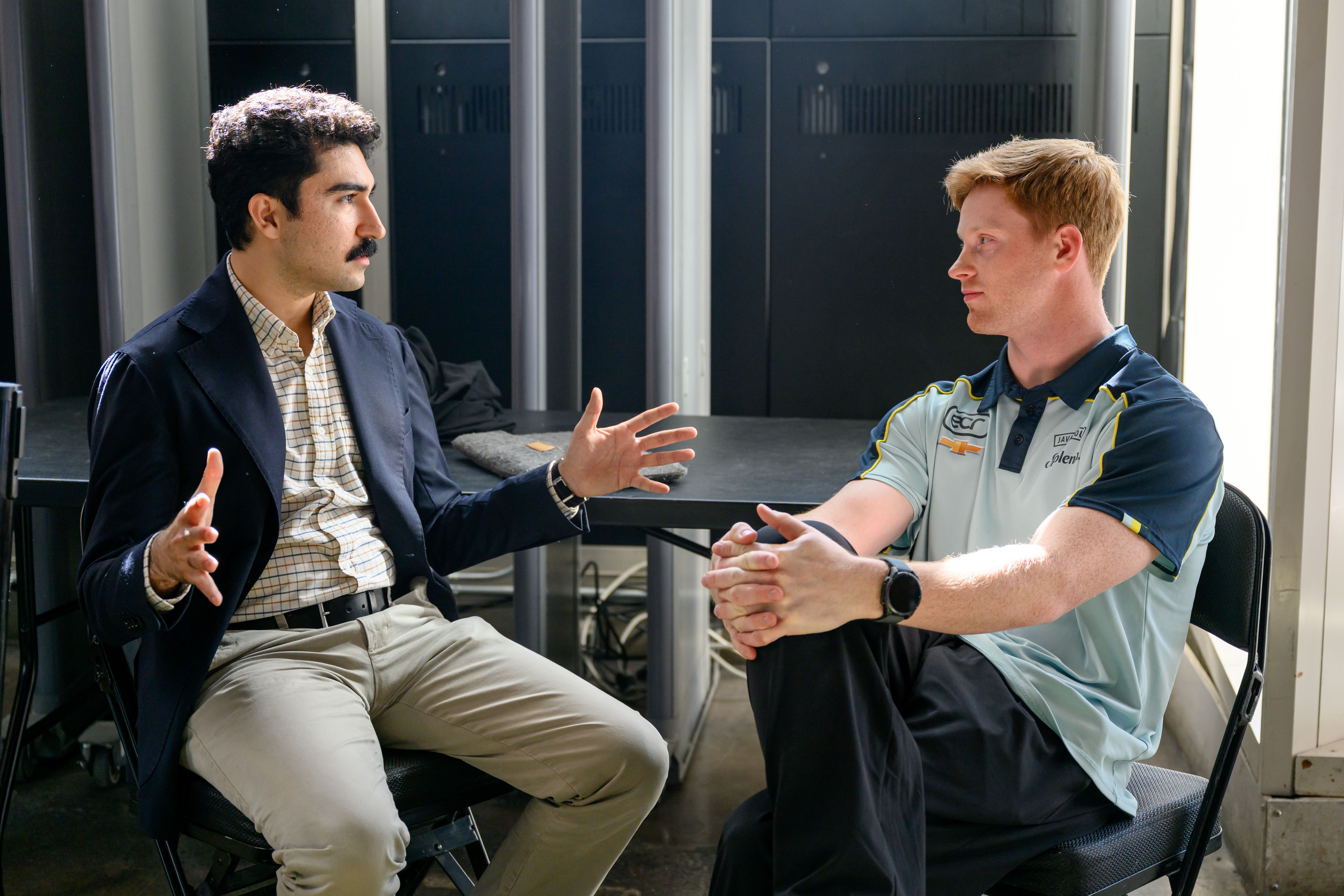 Cuneyt Dil and Christian Rasmussen sit across a table in a server room. The left man (Cuneyt), with a mustache and blazer, gestures with his hands; the right man (Christian) in a light blue polo listens with clasped hands.