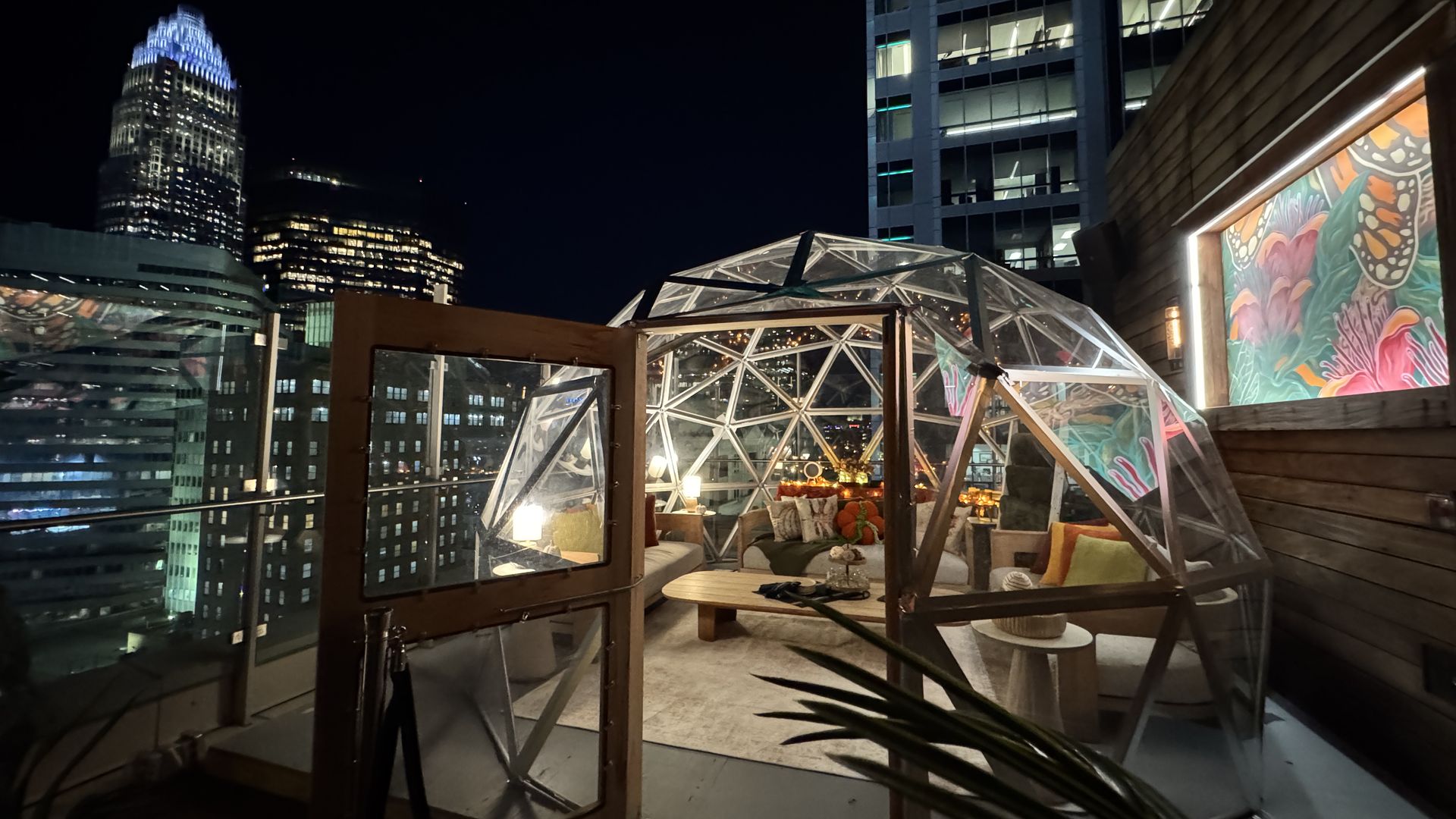 Nighttime rooftop scene with a transparent geodesic dome containing cozy seating, pillows, and warm lighting, city skyscrapers and a colorful framed mural visible nearby.