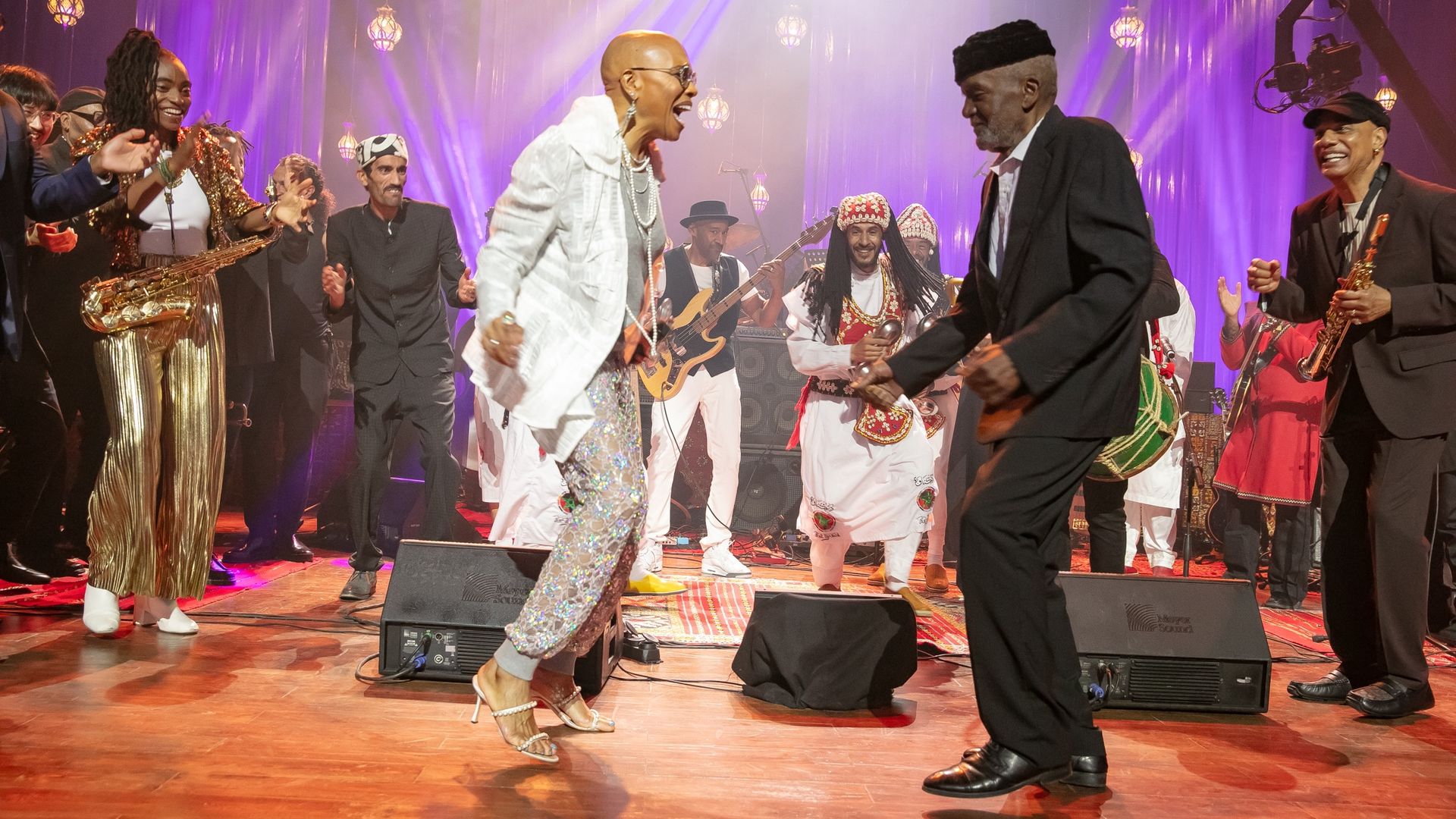 Group of diverse people dancing and playing instruments on stage with purple lighting; woman in white jacket and silver pants and man in black suit dance joyfully center stage.