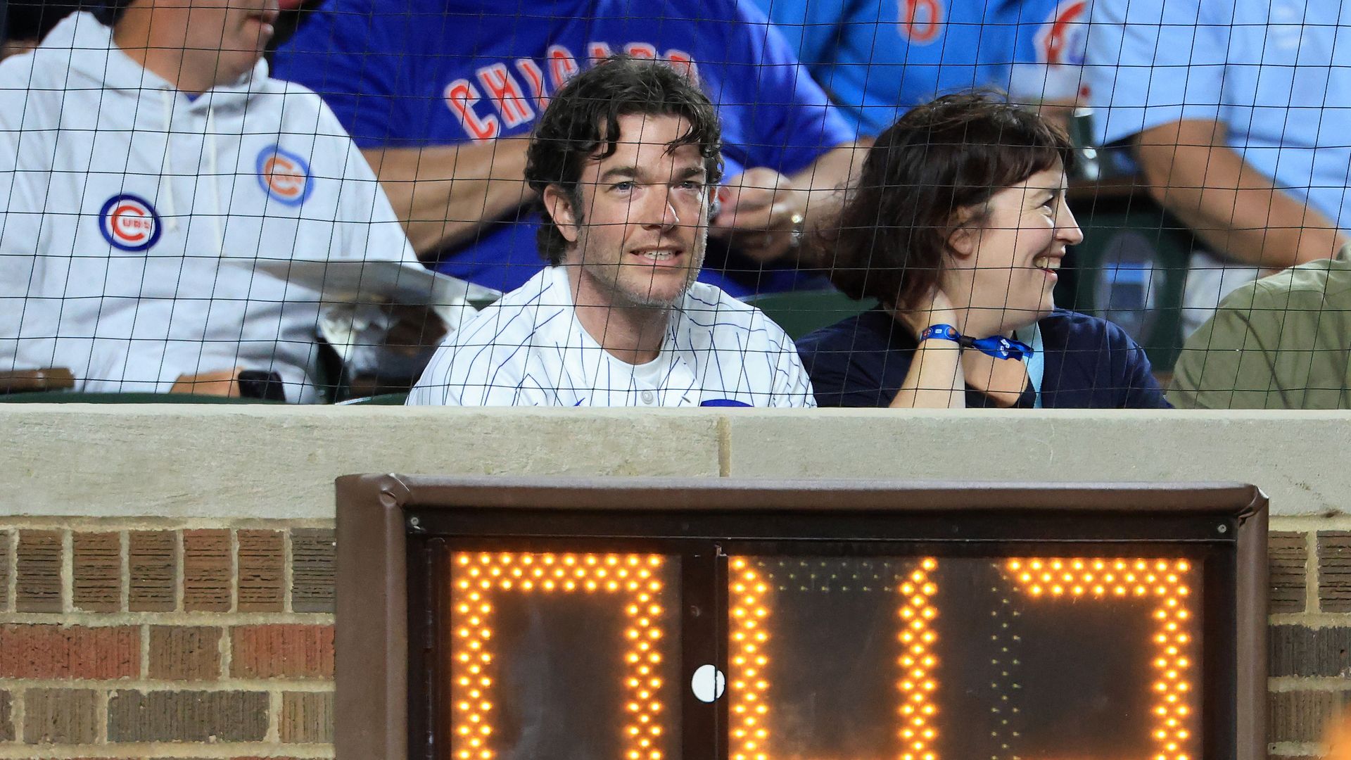 John Mulaney wearing a Cubs jersey sitting behind a fence behind a scoreboard at a baseball game surrounded by people wearing Cubs shirts.