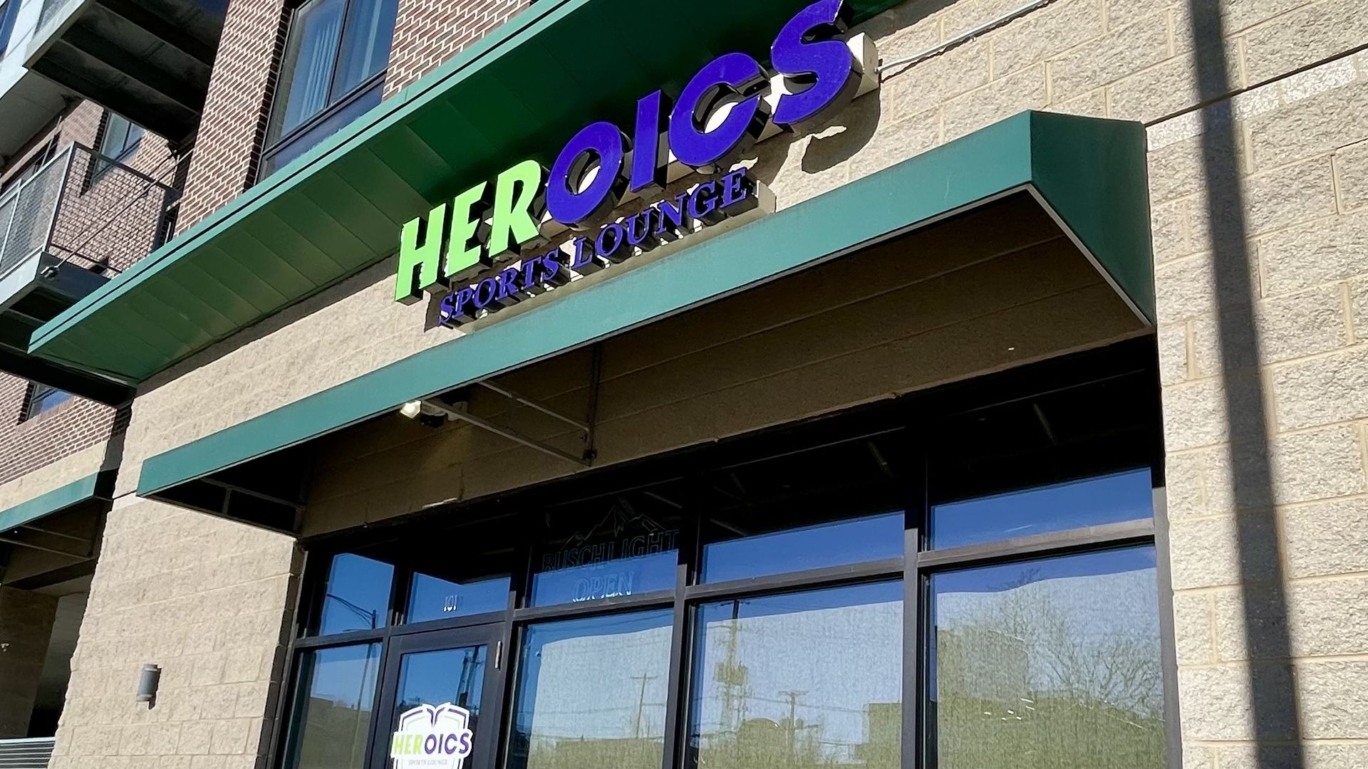 Building entrance with glass doors and windows reflecting the sky, beige stone walls, green awning, and a bright sign reading HEROICS in green and purple, SPORTS LOUNGE below.