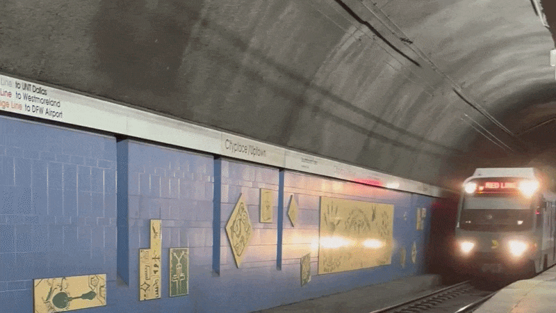 Inside a tunnel, a subway train with headlights approaches on tracks. Blue tiled walls line the platform with white signs and yellow posters; the front displays "RED LINE" in red lights.
