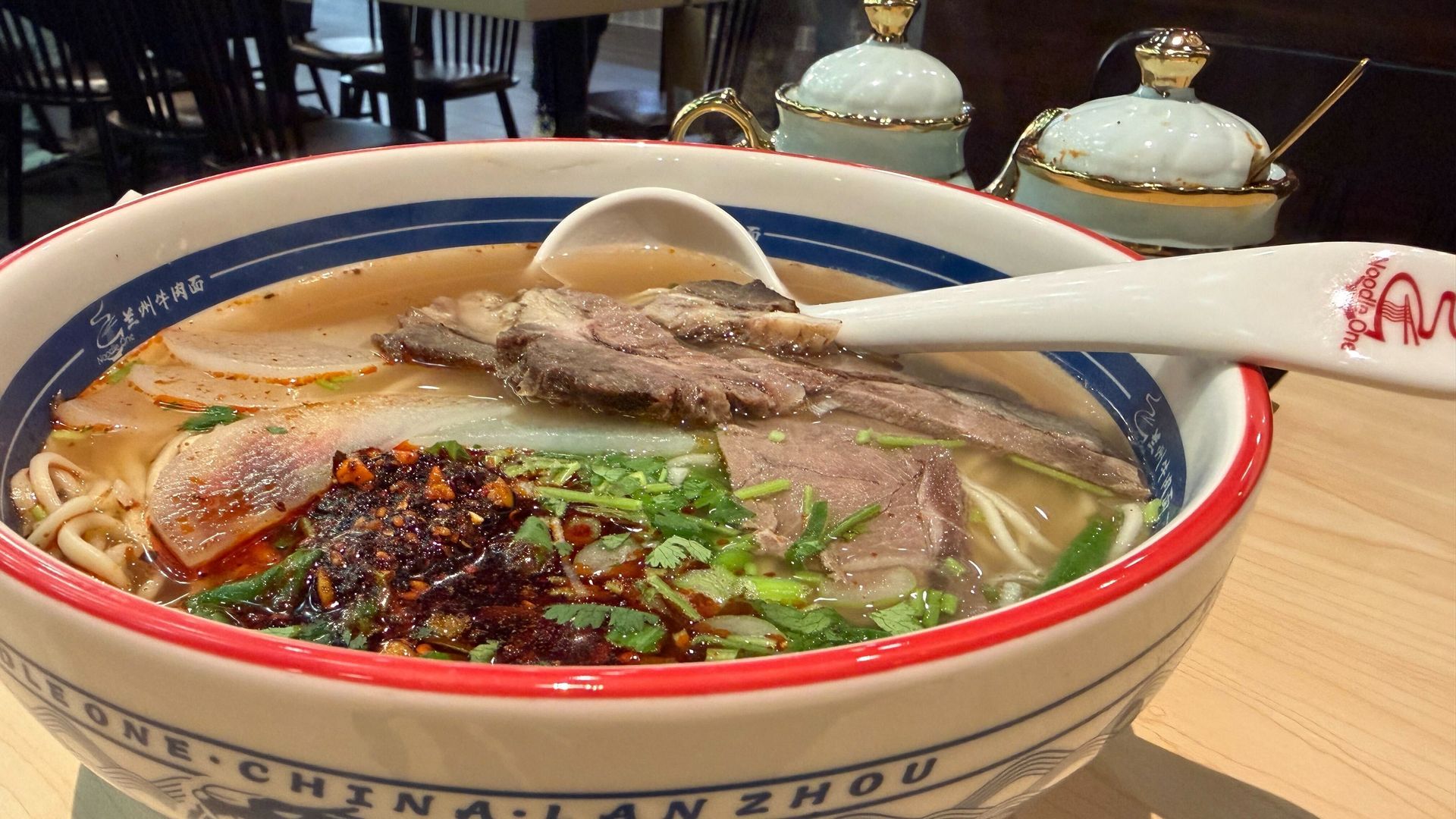 A bowl of noodle soup with slices of beef, chili oil, green herbs, and noodles in a clear broth, served with a white spoon on a wooden table.