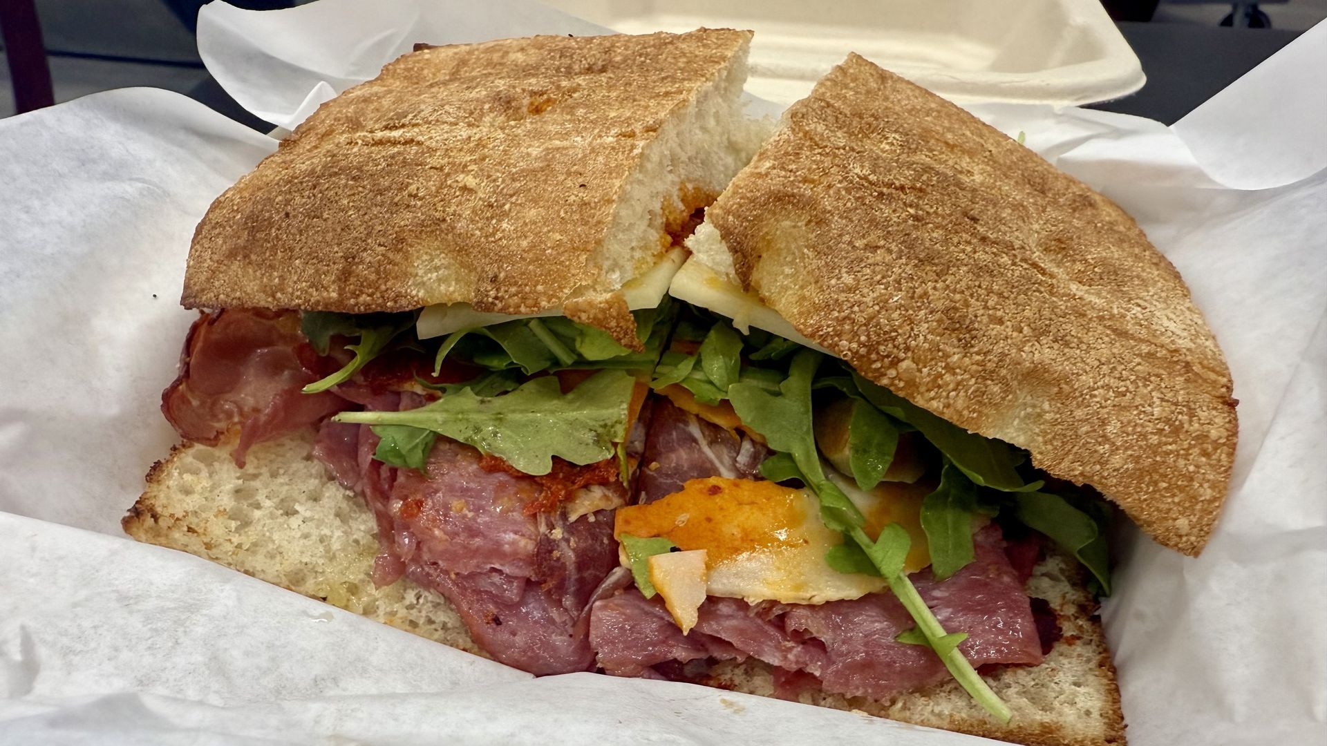 Close-up of a sandwich with toasted crusty bread, layers of pink deli meat, green leafy arugula, and white cheese on white paper.
