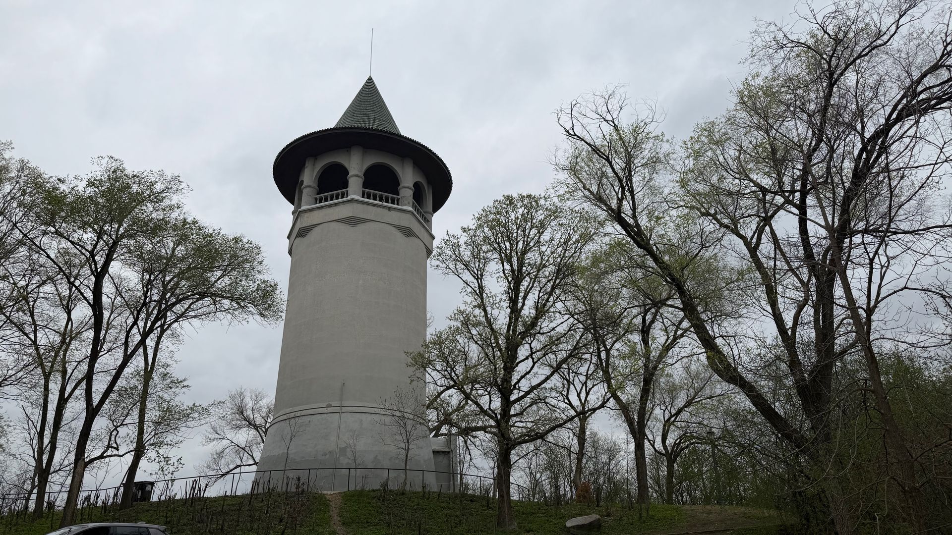 Tall cylindrical concrete tower with a green cone-shaped roof and arched open gallery, surrounded by leafless trees under a gray, overcast sky.
