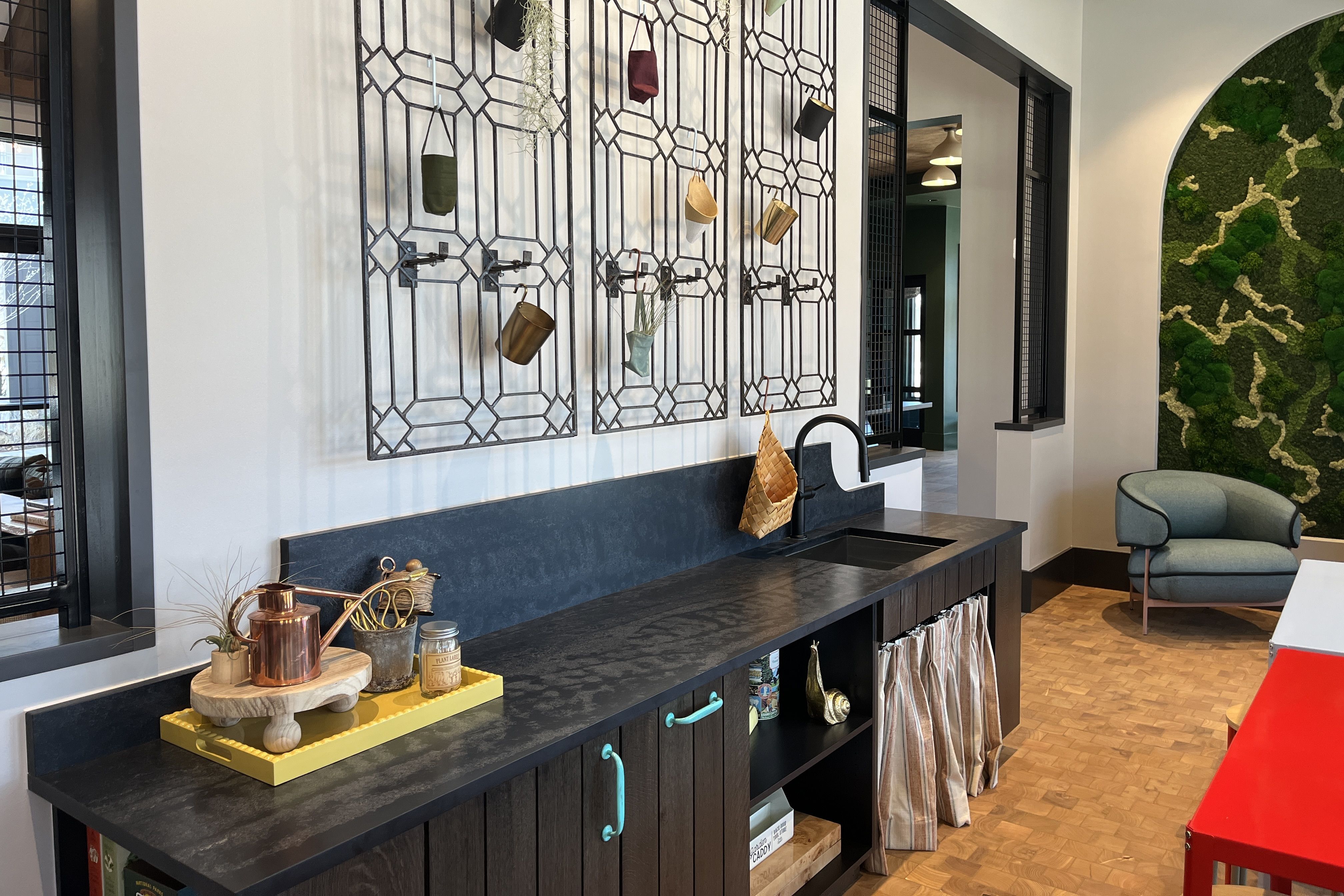 Modern kitchen nook with a dark counter, sink, and turquoise cabinet handles. A geometric metal grid wall holds hanging mugs; copper kettle on a yellow tray. Moss wall and a blue chair nearby.