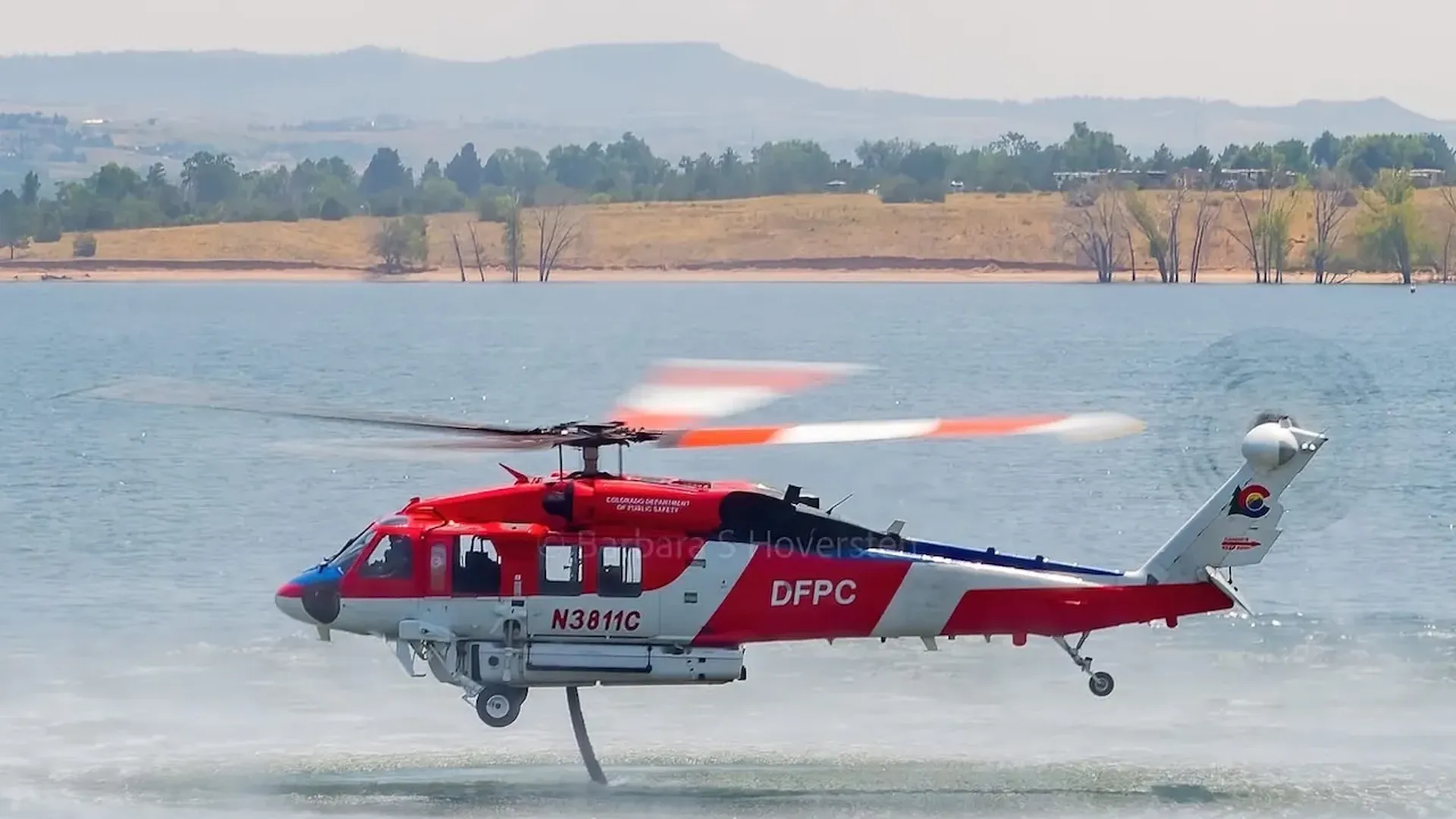 Red-and-white DFPC helicopter with tail number N3811C lifts off from a calm lake; rotor blur and water spray trail behind as shoreline, trees, and distant hills loom in the background.