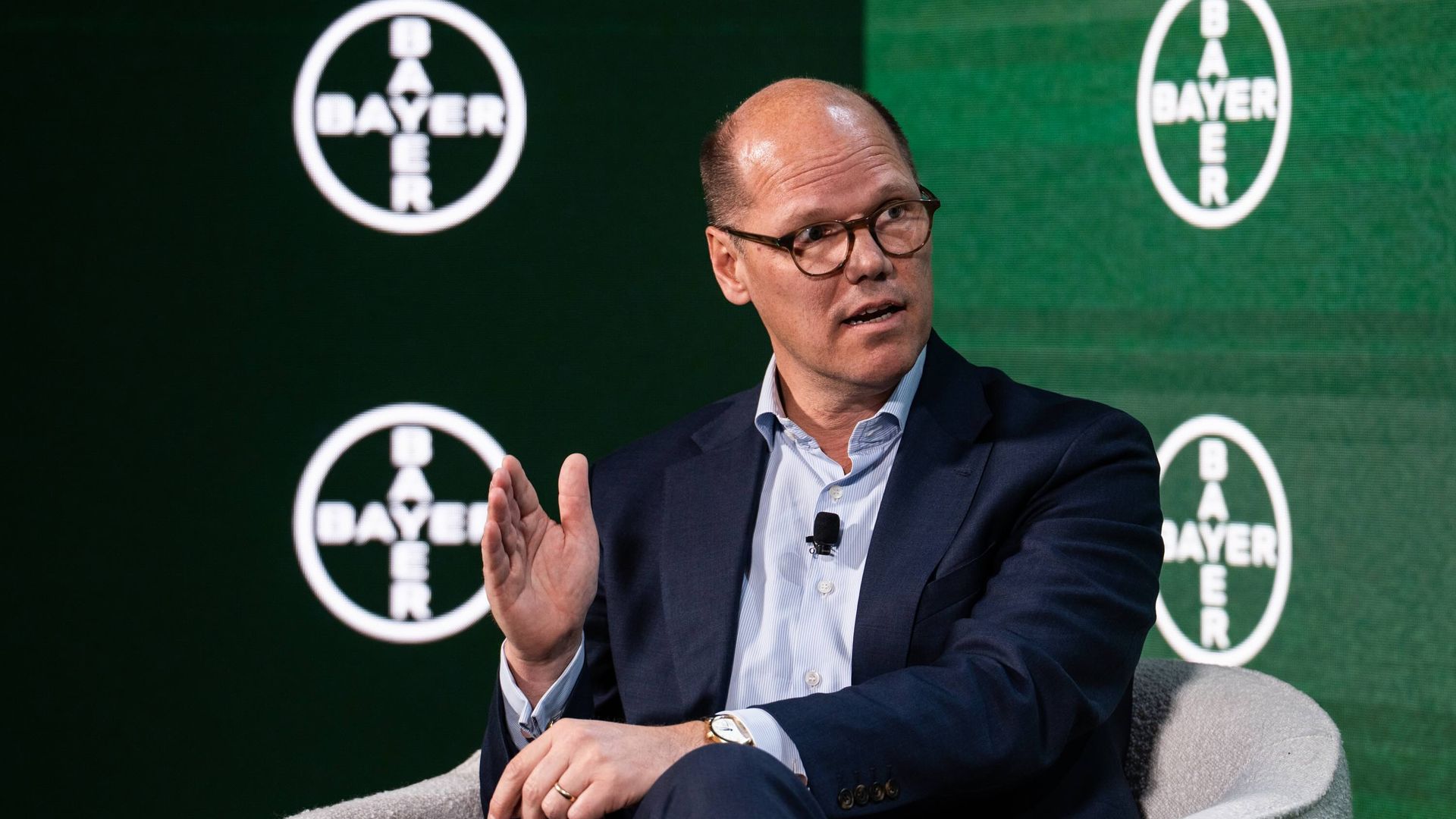 Man in glasses and suit speaking while seated on white chair with multiple green Bayer logos on screen behind him.