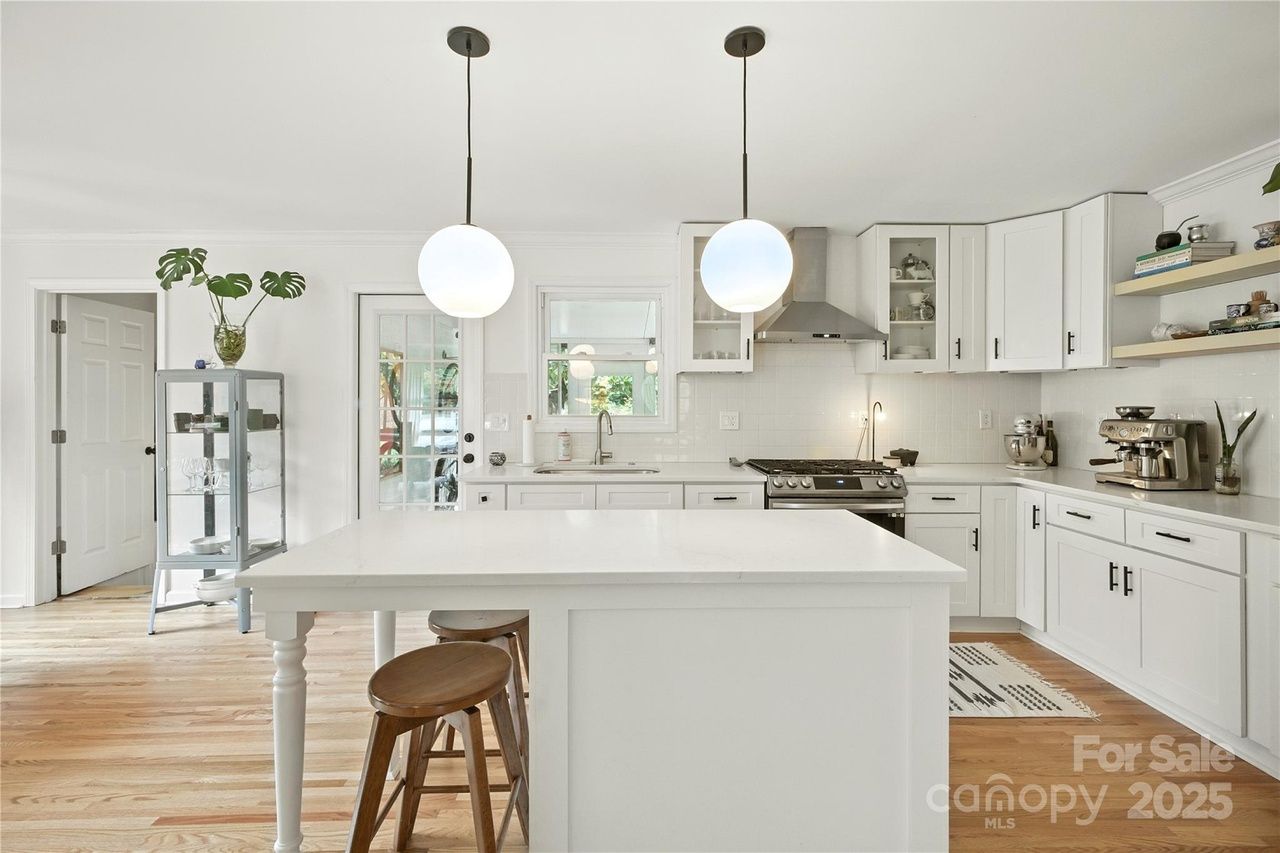 Bright white kitchen with large island, two wooden stools, hanging globe lights, stainless steel stove, glass-front cabinets, and wooden floor with a display cabinet and plants.